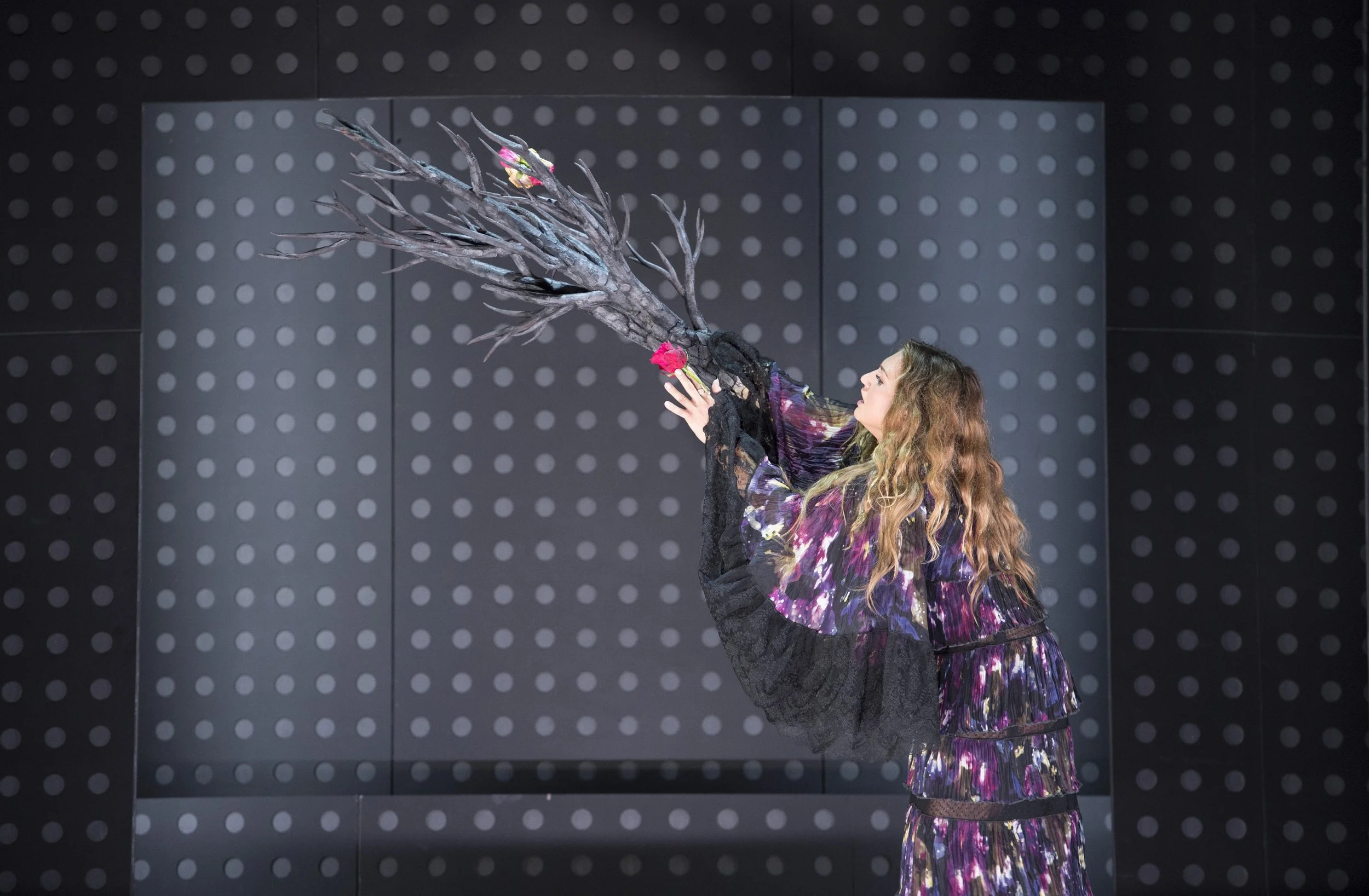 Frau mit langem, gewelltem Haar trägt ein bunt gemustertes Kleid und hält einen künstlichen Baum mit rosa Blumen davor.