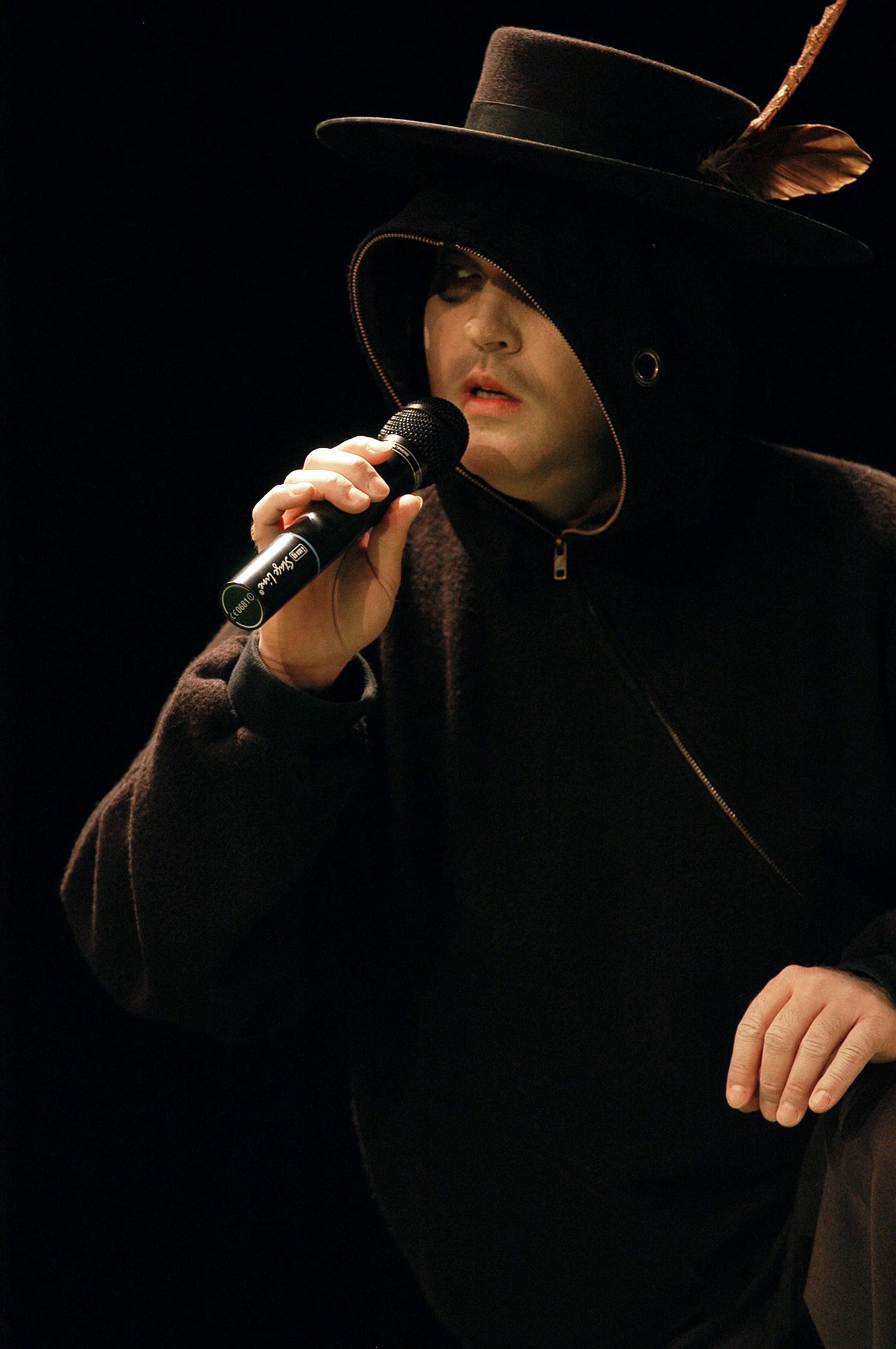 Ein Musiker trägt einen schwarzen Hut mit Feder und einen schwarzen Mantel, während er in ein Mikrofon singt oder spricht.