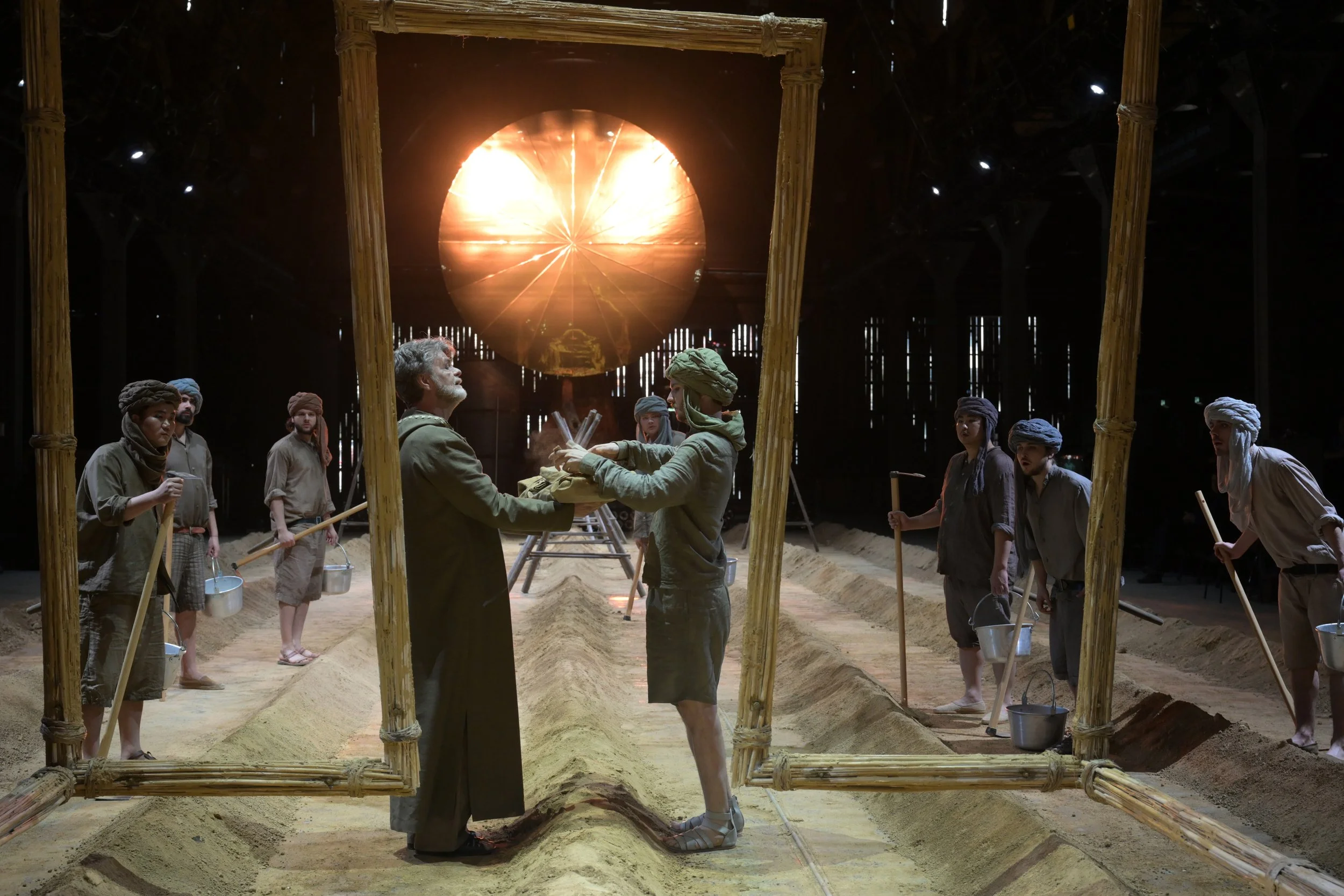 Eine Theater- oder Museumsinszenierung, die eine historische Szene zeigt: Mehrere Figuren in traditionellen Kleidung, die in einer Erde und Holzrahmen befinden, mit einer großen, leuchtenden Sonne an der Rückwand.