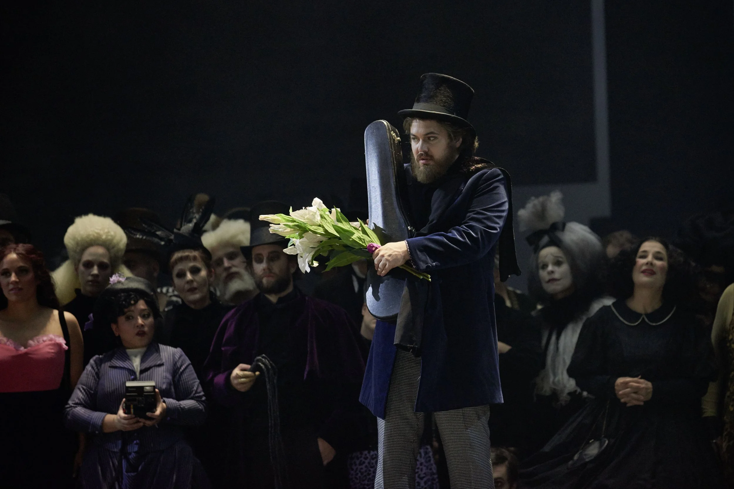 Ein Mann mit Zylinderhut hält eine Blumenstrauß in einer schwarzen Tasche in der Hand, umgeben von anderen Personen in Kostümen, wahrscheinlich bei einer Theateraufführung oder einem Kostümball.