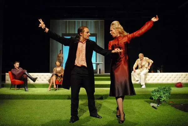 Eine Theaterversammlung mit fünf Schauspielern auf einer grünen Bühne. Zwei der Schauspieler im Vordergrund tanzen oder performen, während drei weitere im Hintergrund zuschauen.