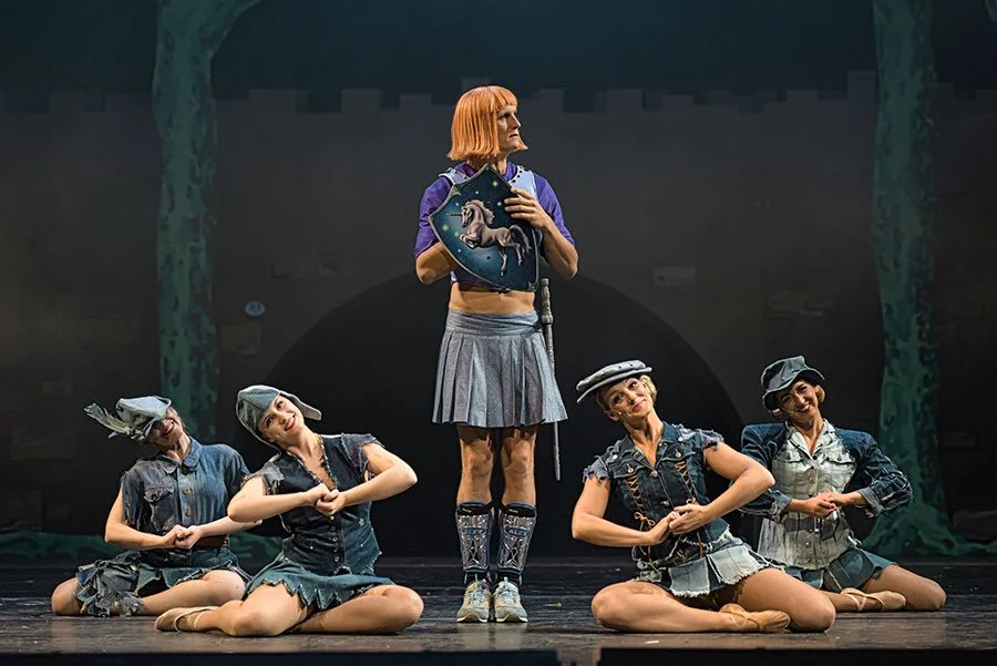 Balletttänzer auf Bühne, eine steht in der Mitte mit einem Schutzschild, die anderen sitzen im Schneidersitz und posieren.