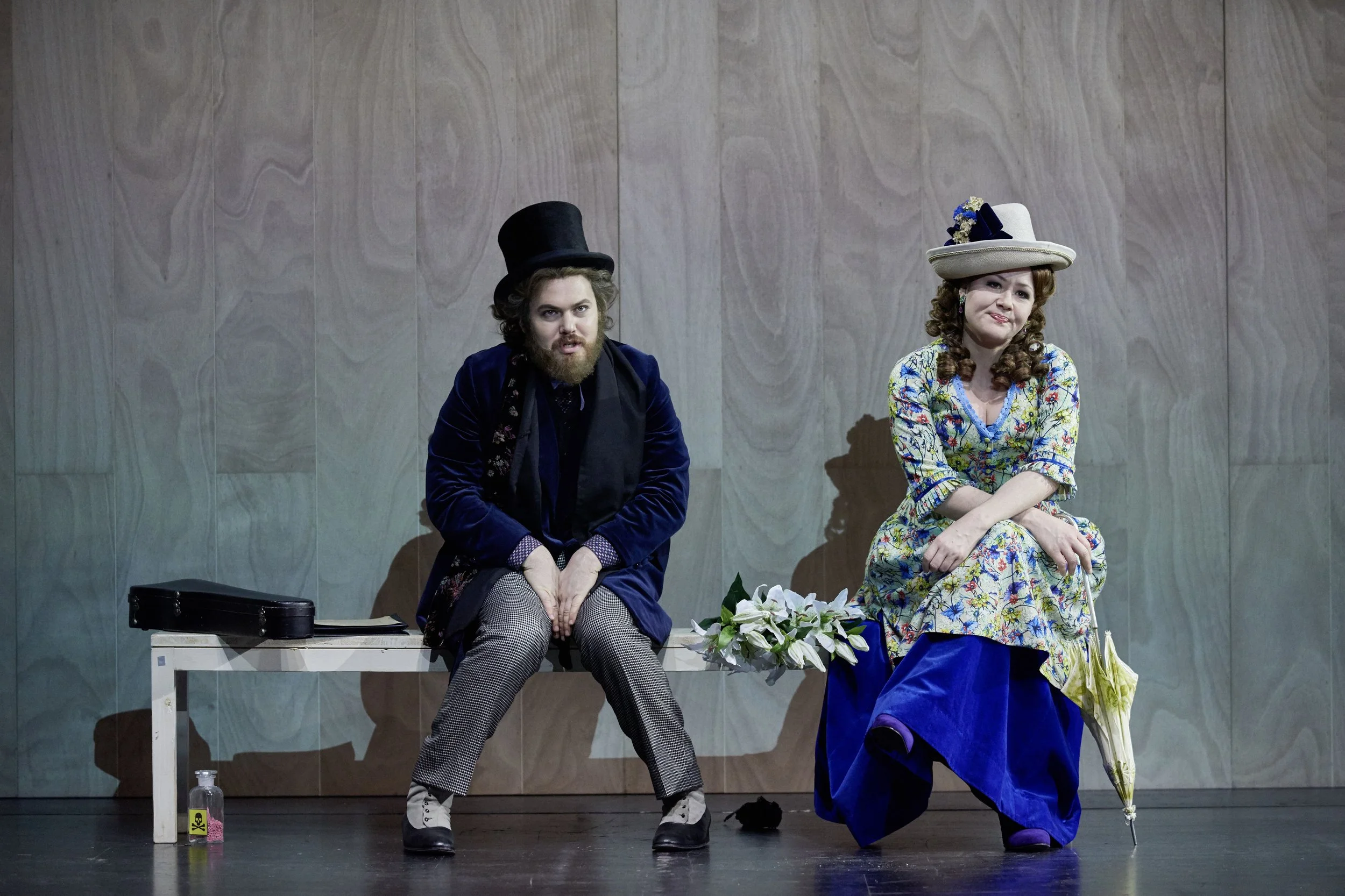 Ein Mann und eine Frau sitzen auf einer Bank auf einem Bühnenbild. Der Mann trägt einen Zylinderhut, einen blauen Mantel und karierte Hosen. Die Frau trägt einen Hut mit Blumen, ein geblümtes Kleid und eine blaue Unterrock. Sie hält einen Schirm und 