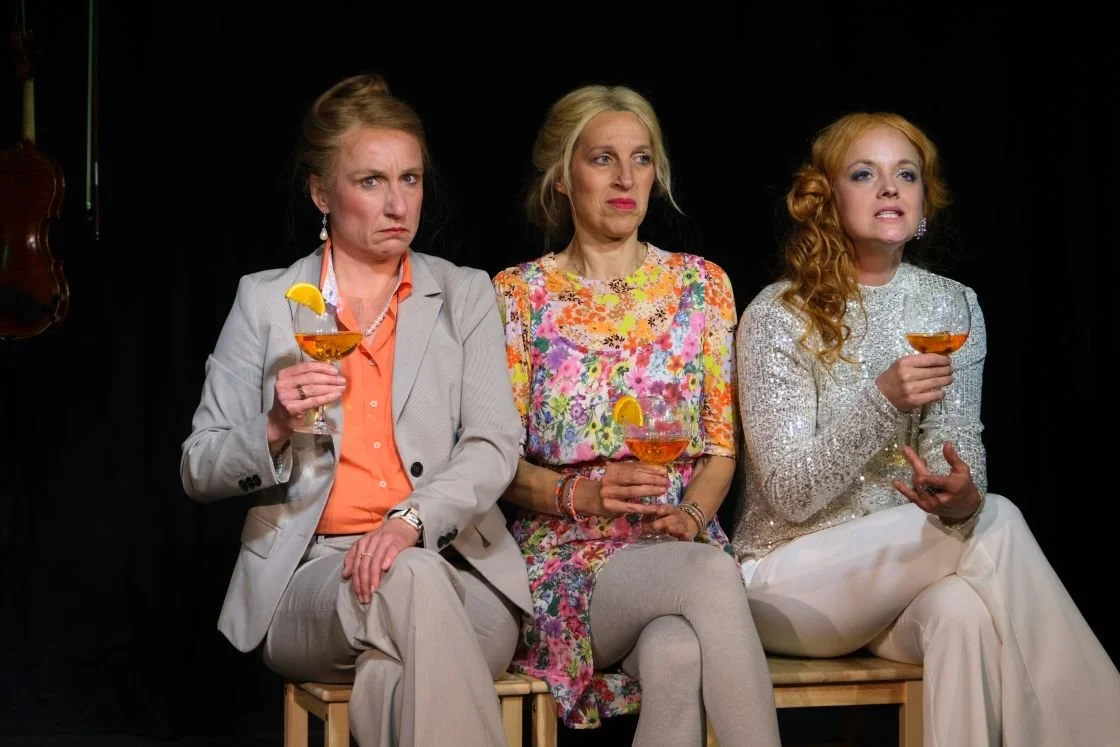 Drei Frauen sitzen nebeneinander auf einer Bank, jede hält einen Cocktail mit einem Zitronenschnitz. Sie wirken ernst und konzentriert.