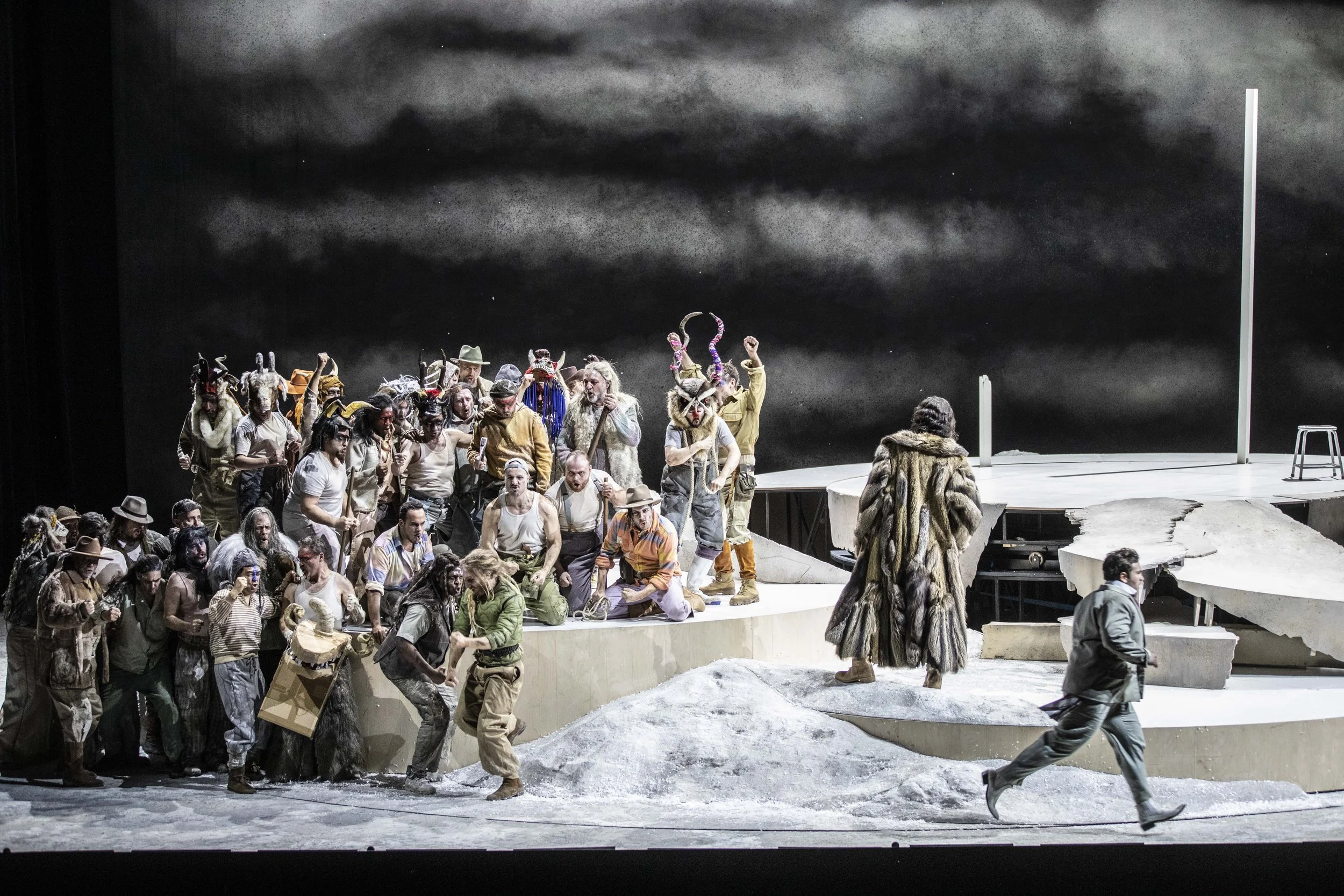 Bühnenbild einer Theaterproduktion mit mehreren Schauspielern in Tierkostümen, die auf einer Kulisse mit dunklem, wolkigem Himmel stehen, einige treten auf der provisorischen Bühne, andere bewegen sich im Vordergrund.