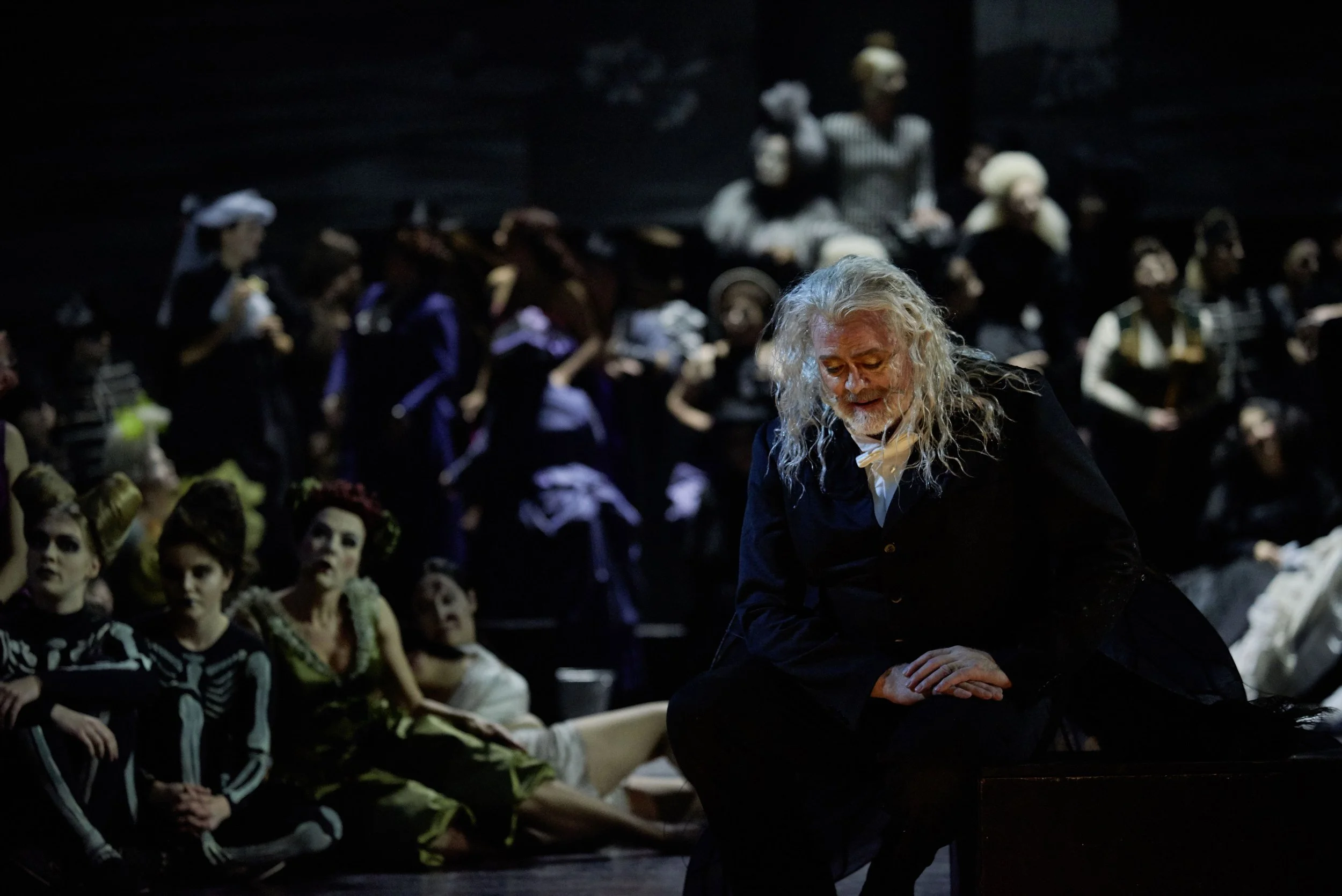 Schauspieler in Kostümen auf einer Bühne während einer Theateraufführung, eine Figur mit weißen, lockigen Haaren im Vordergrund, umgeben von anderen Darstellern in dunkler und bunter Kleidung.