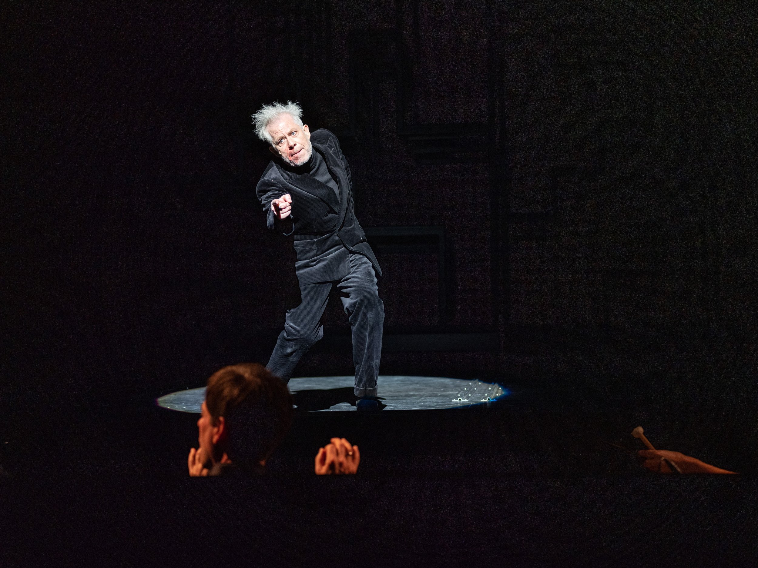 Theateraufführung mit einem älteren Mann, der auf der Bühne im Licht steht und eine Figur spielt, während das Publikum zusieht.