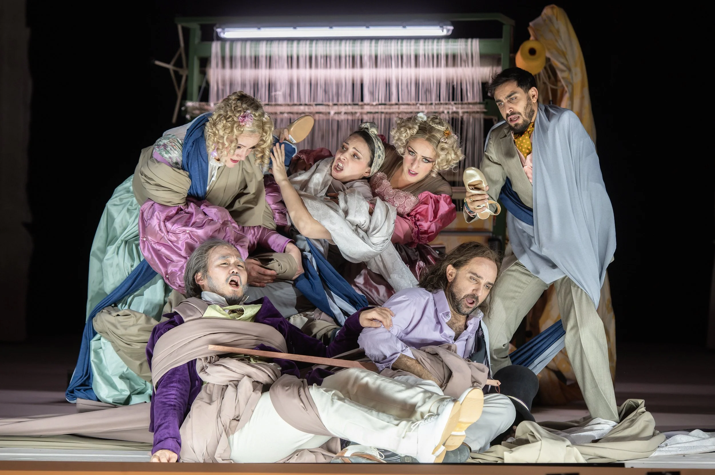 Szenenfoto einer Theatervorführung mit mehreren Schauspielern in farbenfrohen, historischen Kostümen, die auf einer Bühne eine dramatische Szene darstellen, mit einem grünen Kulissenrahmen im Hintergrund.