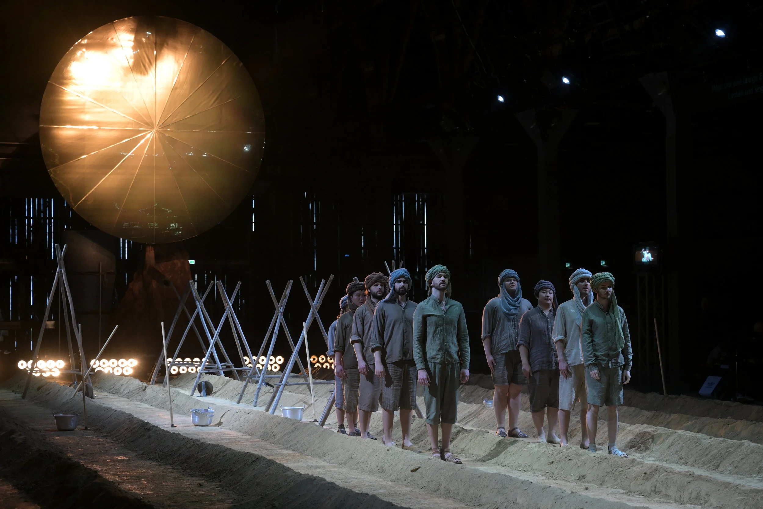 Theaterstück mit mehreren Darstellern in einem dunklen Studio, Sandboden und einer großen Goldkugel im Hintergrund.