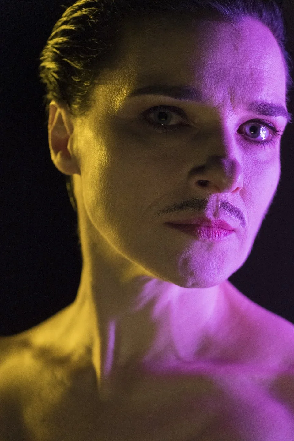 Porträt eines Mannes mit leichtem Bart, beleuchtet von buntem Licht in Pink und Gelb, gegen schwarzen Hintergrund.