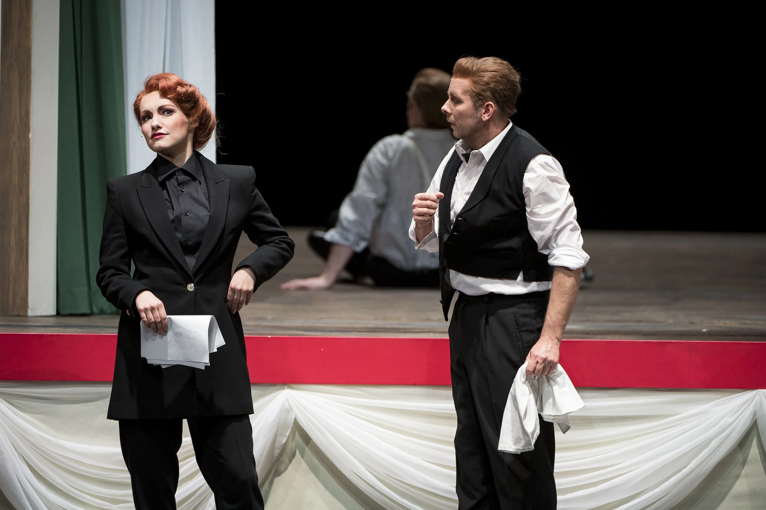 Zwei Schauspieler auf einer Theaterbühne, eine Frau mit roten Haaren in einem schwarzen Anzug und eine Mann in einem weißen Hemd, schwarzer Weste, im Gespräch, im Hintergrund eine weitere Person sitzend auf der Bühne.