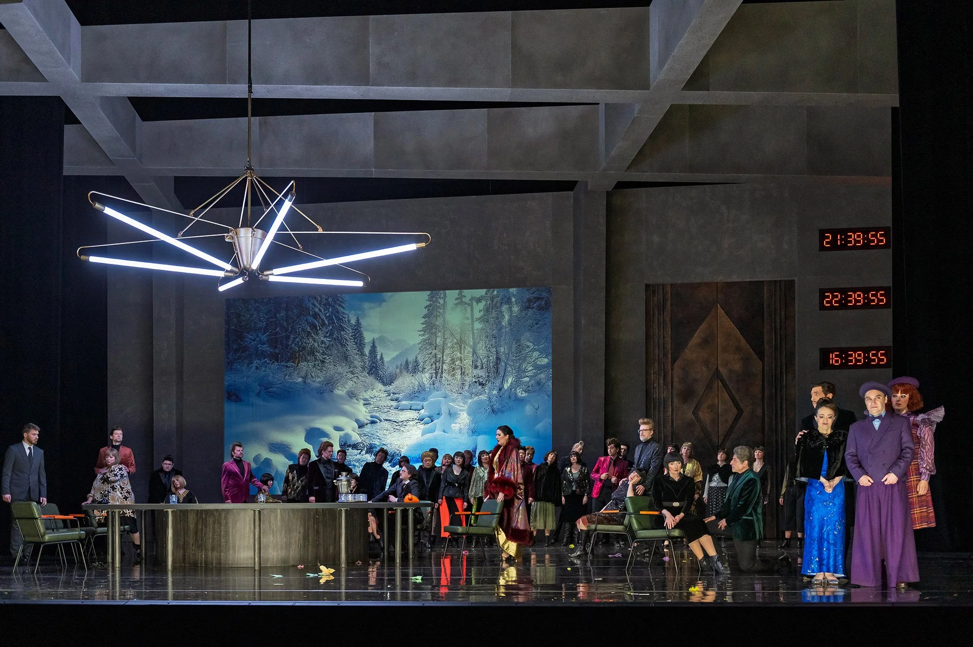 Bühnenbild einer Theaterproduktion mit mehreren Schauspielern, moderner und bunter Kostüme, digitaler Uhr und Winterlandschaft im Hintergrund.