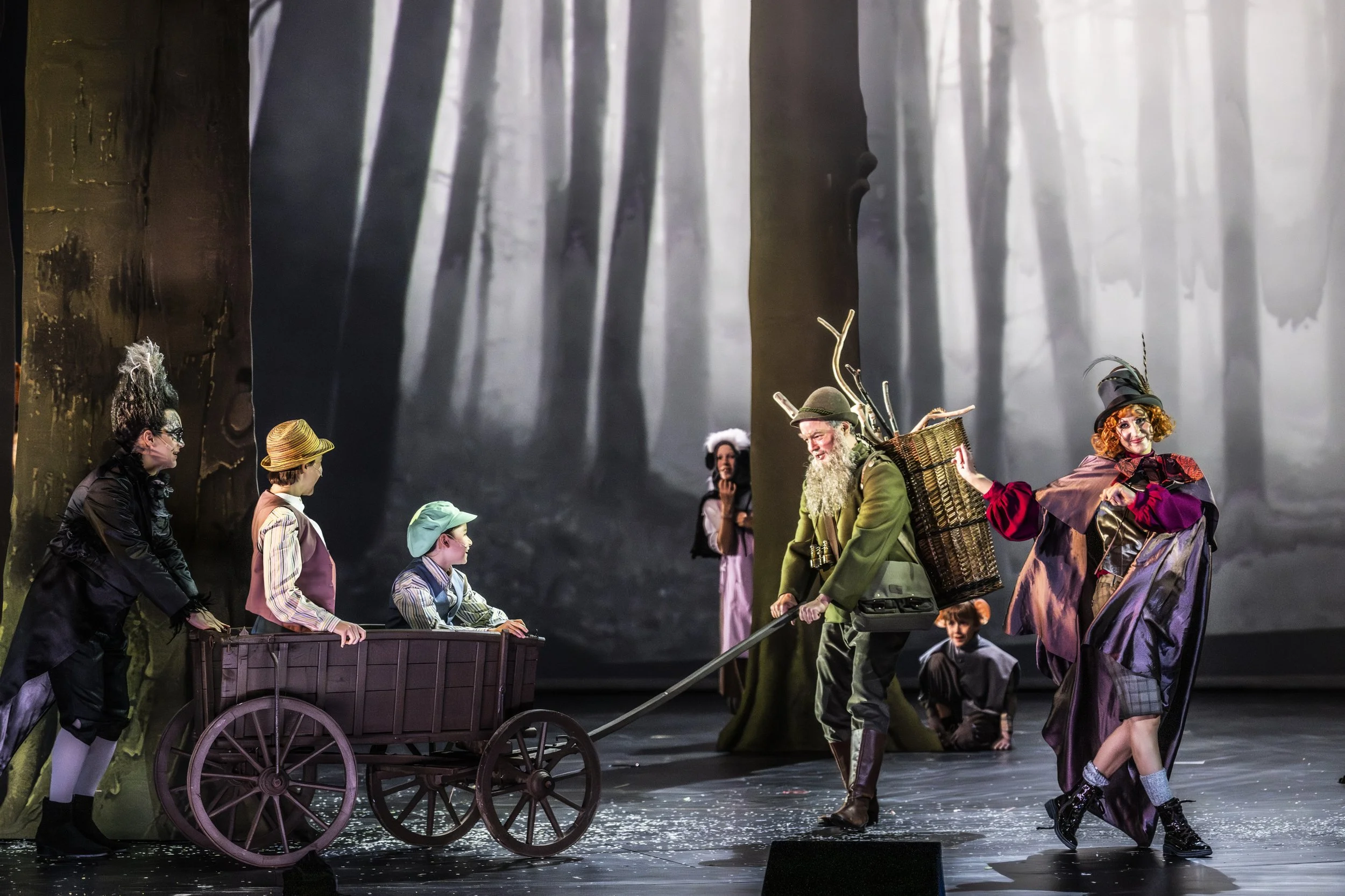 Szenen aus einem Theaterstück mit mehreren Schauspielern in einem Waldsetting, darunter Kinder in einem Holzwagen, einen alten Mann mit Stock und Rucksack, eine Frau in bunter Kleidung und andere Figuren in periodischer Kleidung.