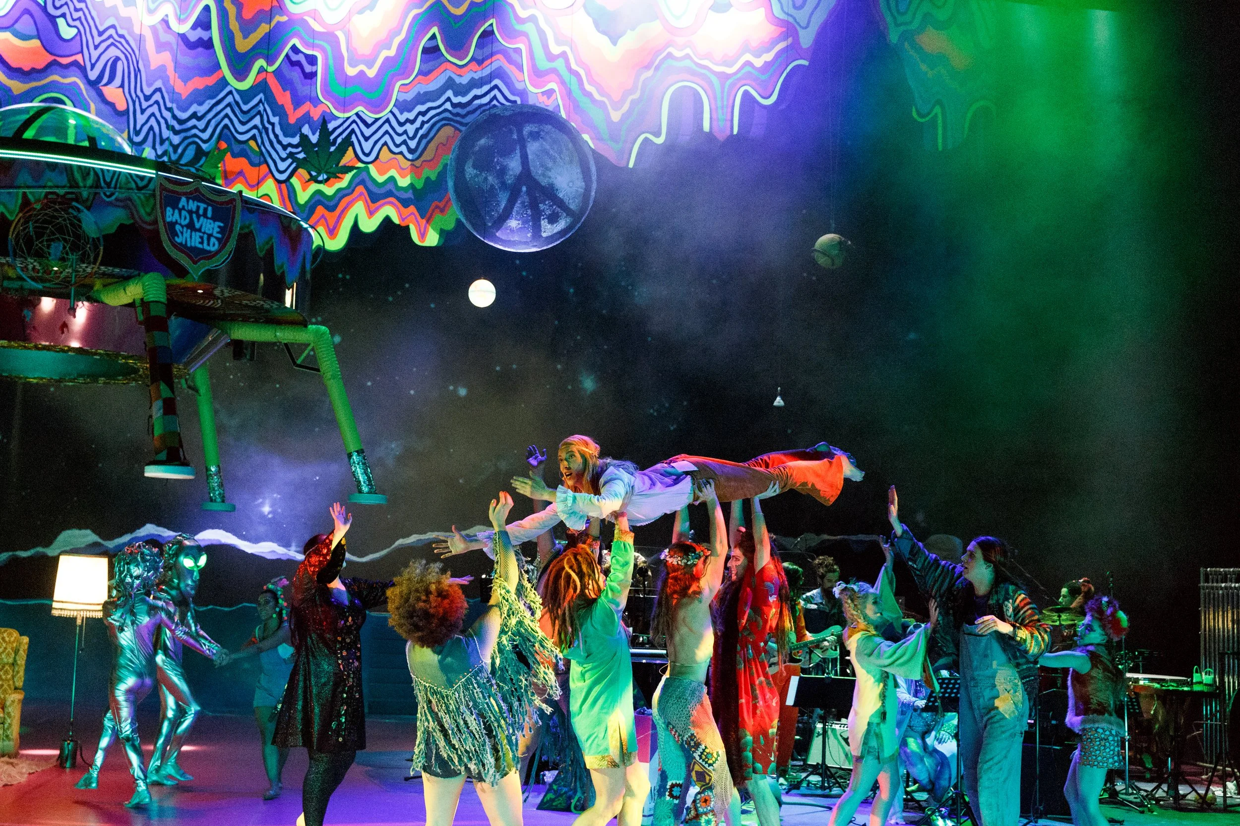 Menschen, die bei einer bunten Theateraufführung auf einer Bühne tanzen, mit einer Frau, die wie Jesus dargestellt ist, die auf den Händen anderer liegt, unter einer psychedelischen, farbenfrohen Decke und weißen Sternen im Hintergrund.