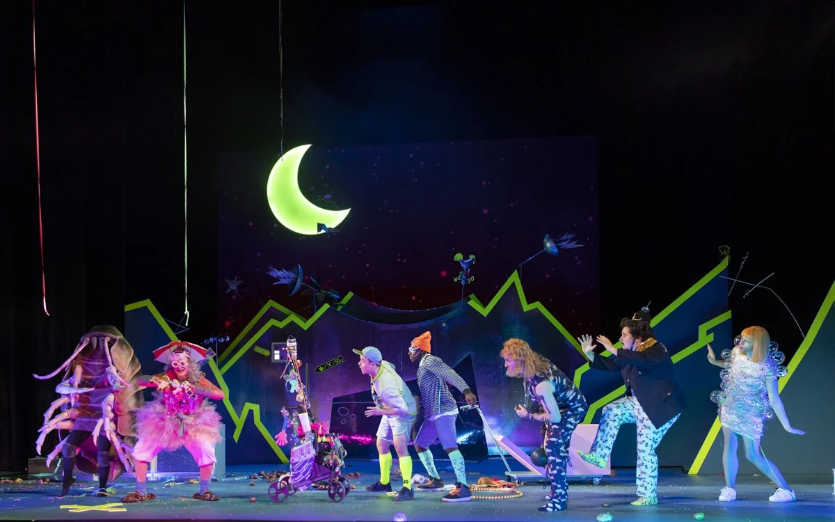 Bühnenaufführung mit mehreren bunt gekleideten Schauspielern, fantasievoller Kulisse mit grünen und blauen Grafiken, Mond und Sternen. Schauspieler in skurrilen Kostümen, einige mit Requisiten wie Regenschirm, Rollstuhl, Glühbirne. Szenen mit Bewegun