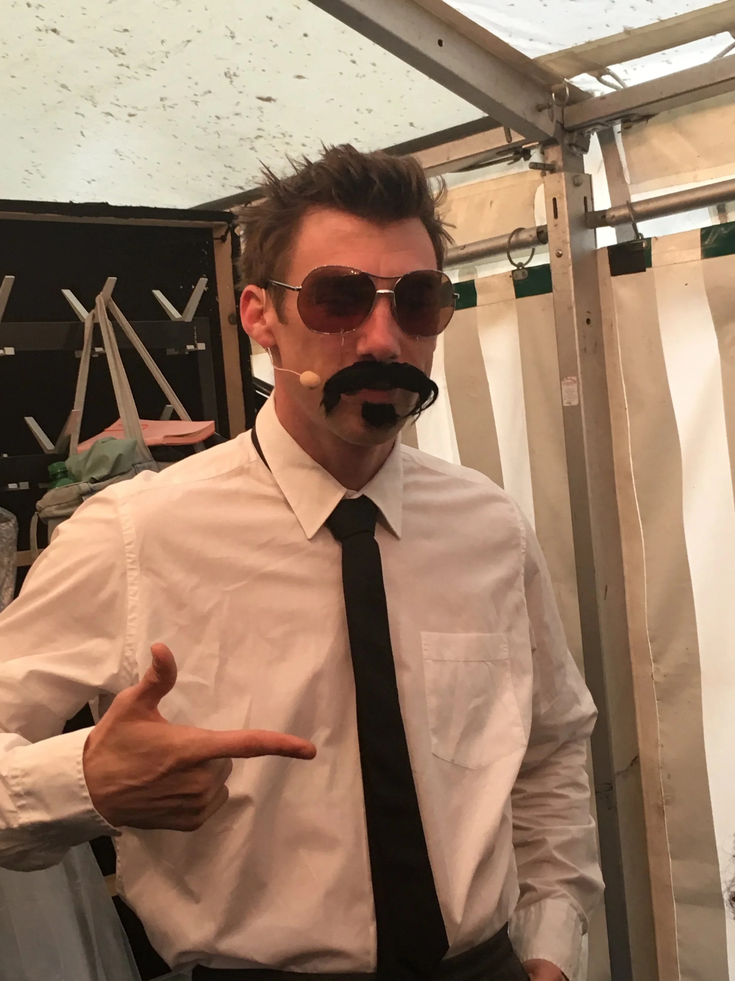 Mann in Verkleidung mit Sonnenbrille, schwarz-weißem Hemd, schwarzer Krawatte, Schnurrbart und Koteletten, zeigt auf sich selbst.