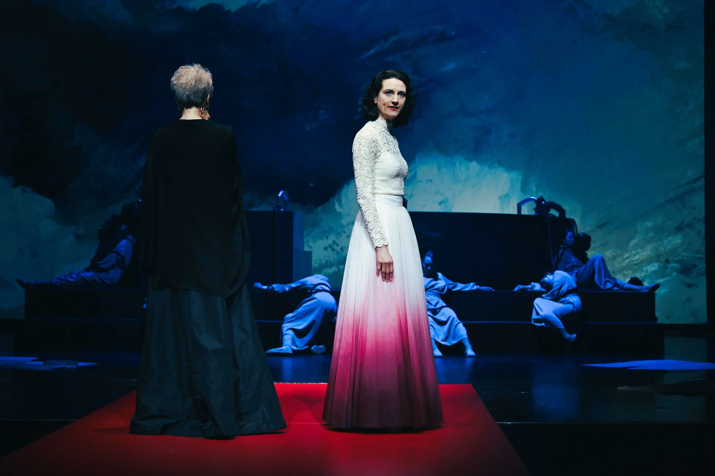 Drei Frauen auf einer Bühne, eine im Vordergrund trägt ein langes, weißes Kleid mit rotem Farbverlauf. Die andere steht mit dem Rücken zum Publikum. Im Hintergrund sind Männer in weißen Gewändern, die auf einer Plattform liegen.