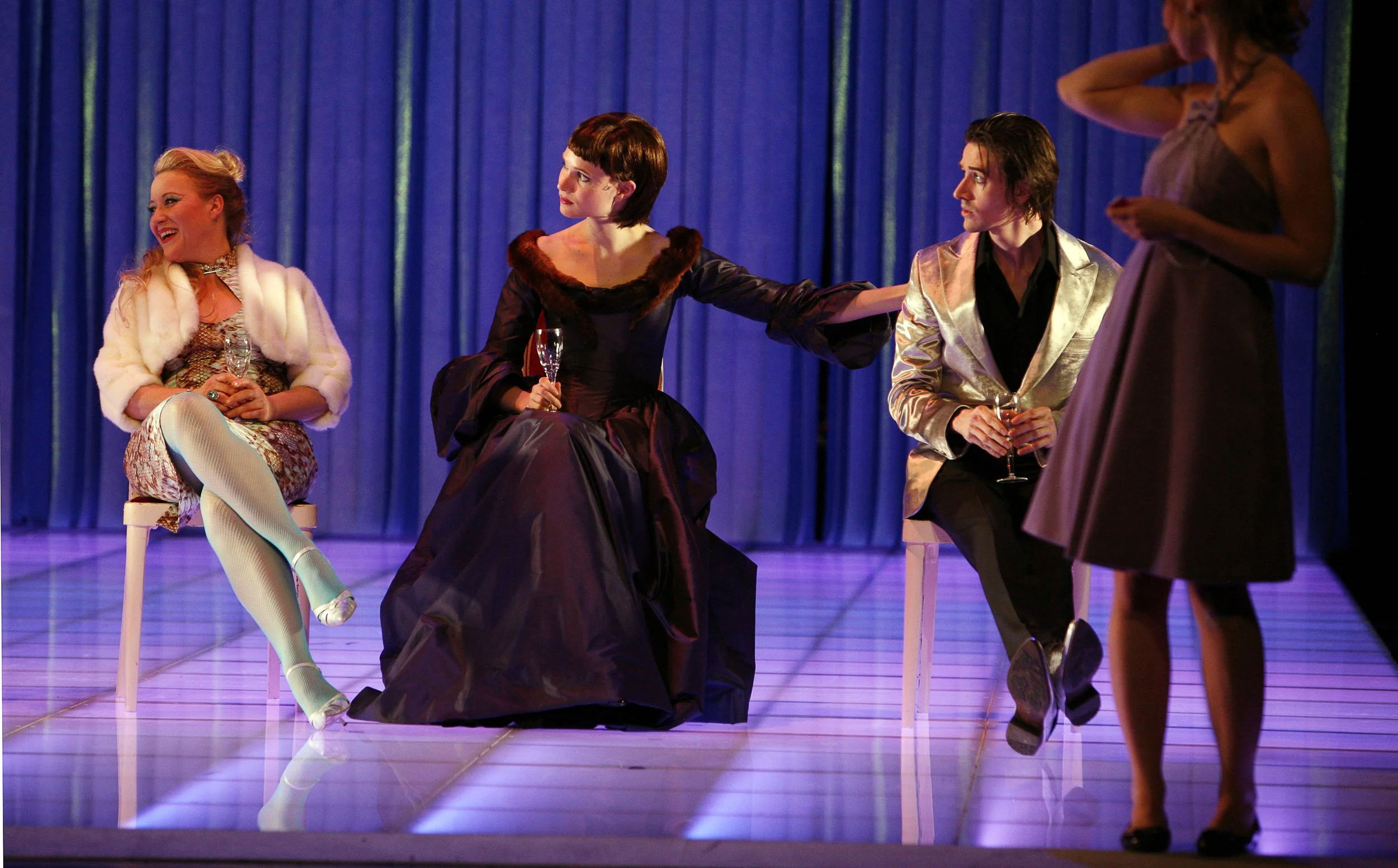 Vier Schauspielerinnen auf einer Bühne mit blauer Vorhang, die in einem Theaterstück eine Szene darstellen, mit Gläsern in den Händen.