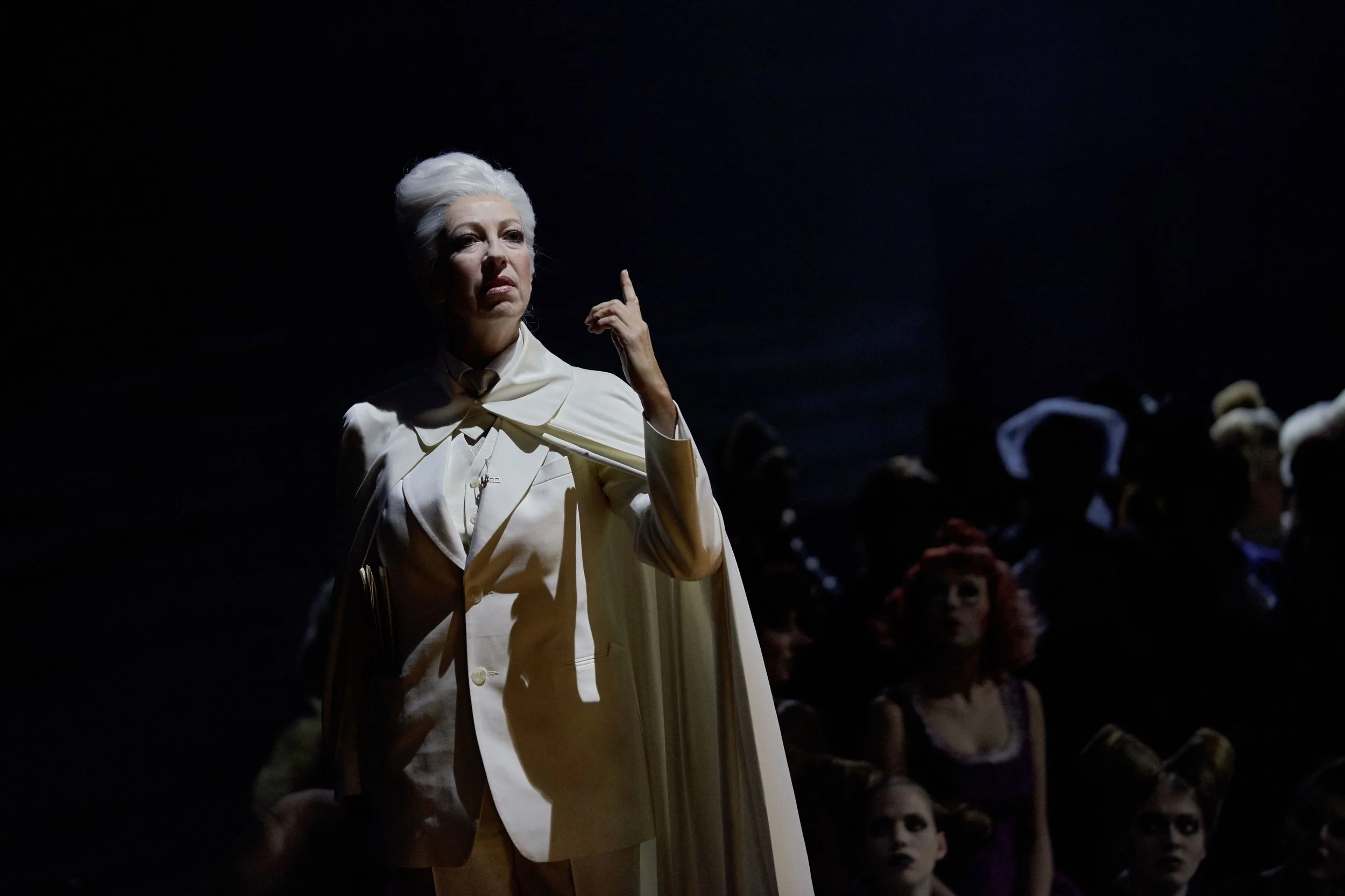 Theateraufführung mit einer Frau in weißem Kostüm, die auf der Bühne steht, und mehreren Publikumsmitgliedern im Schatten im Hintergrund.