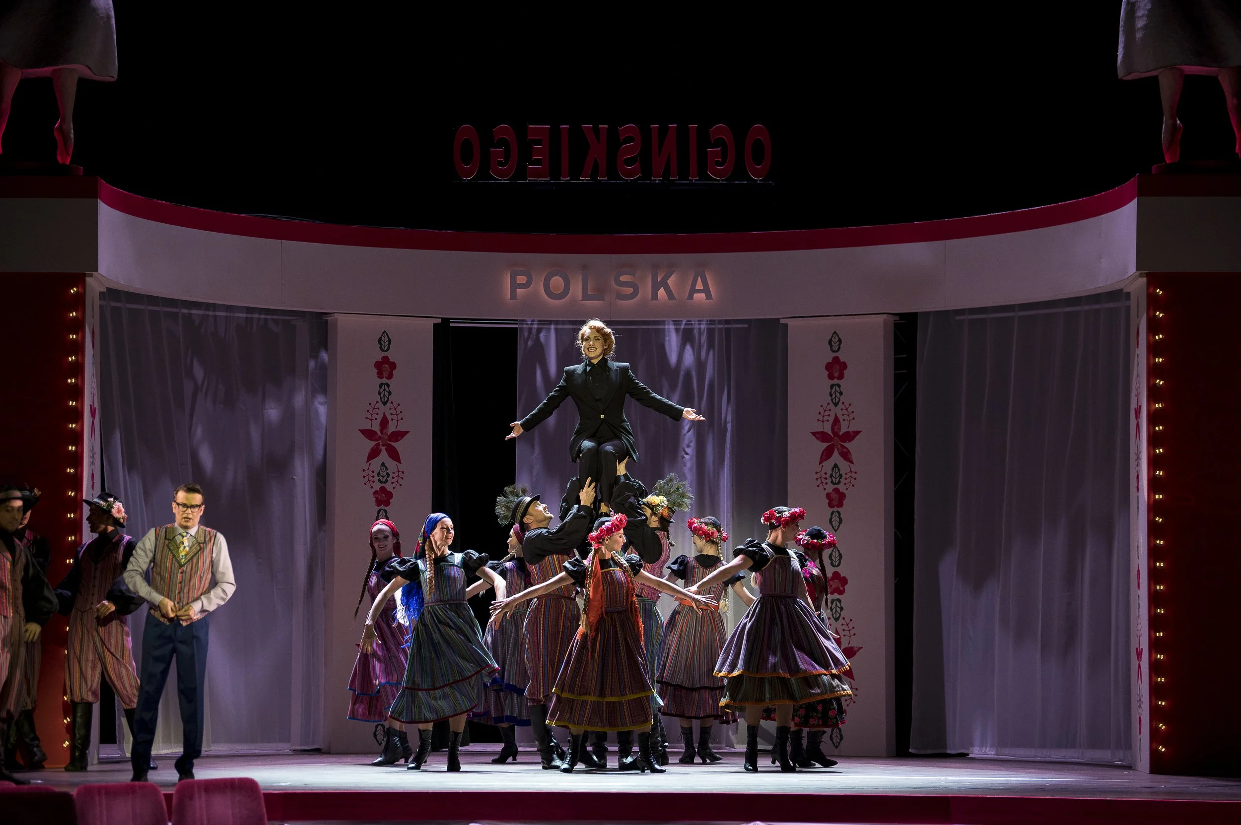Bühnenaufführung mit Tänzern in traditionellen polnischen Trachten und einer Solistin, die auf den Schultern eines Tänzers steht, vor einer Kulisse mit den Worten 'POLSKA' und 'OGINSKIEGO'.