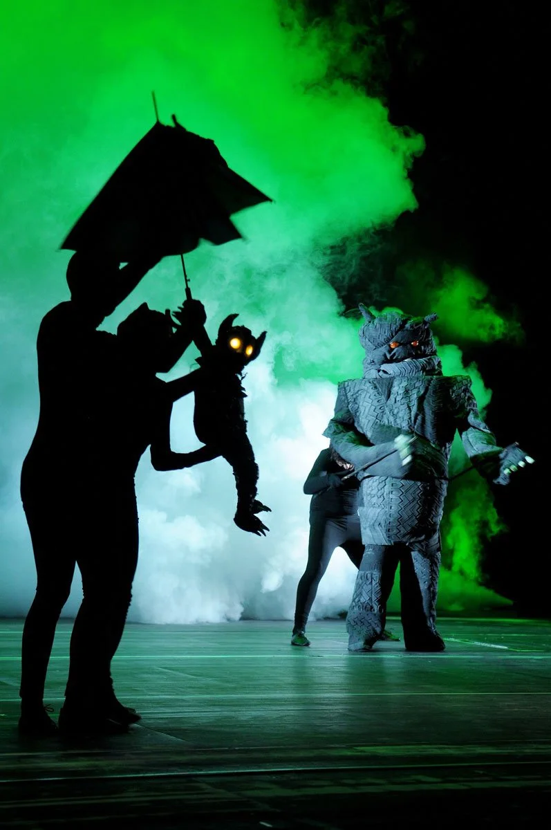 Theaterbühne mit einem Menschen in einem Krokodilmaskottkostüm und einer anderen Person in einem schwarzen Kostüm mit leuchtenden Augen, die an einem Requisit hängt, bei grünem Rauch im Hintergrund.