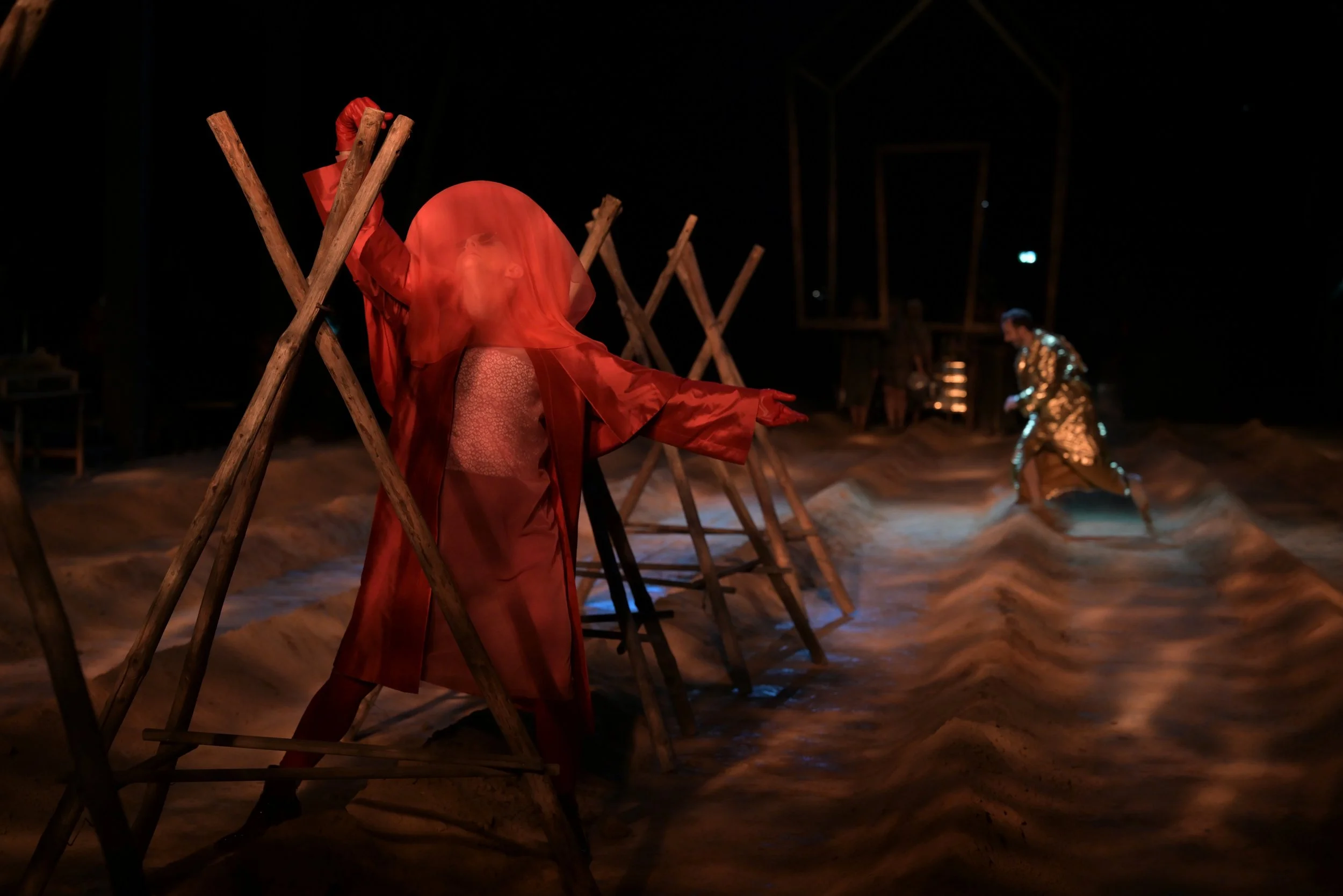 Drei Menschen in künstlerischer Performance, eine in rot mit einem transparenten Schleier, stehend zwischen Holzgerüsten, andere in schimmernden Kleidern im Sandstadion bei Nacht.