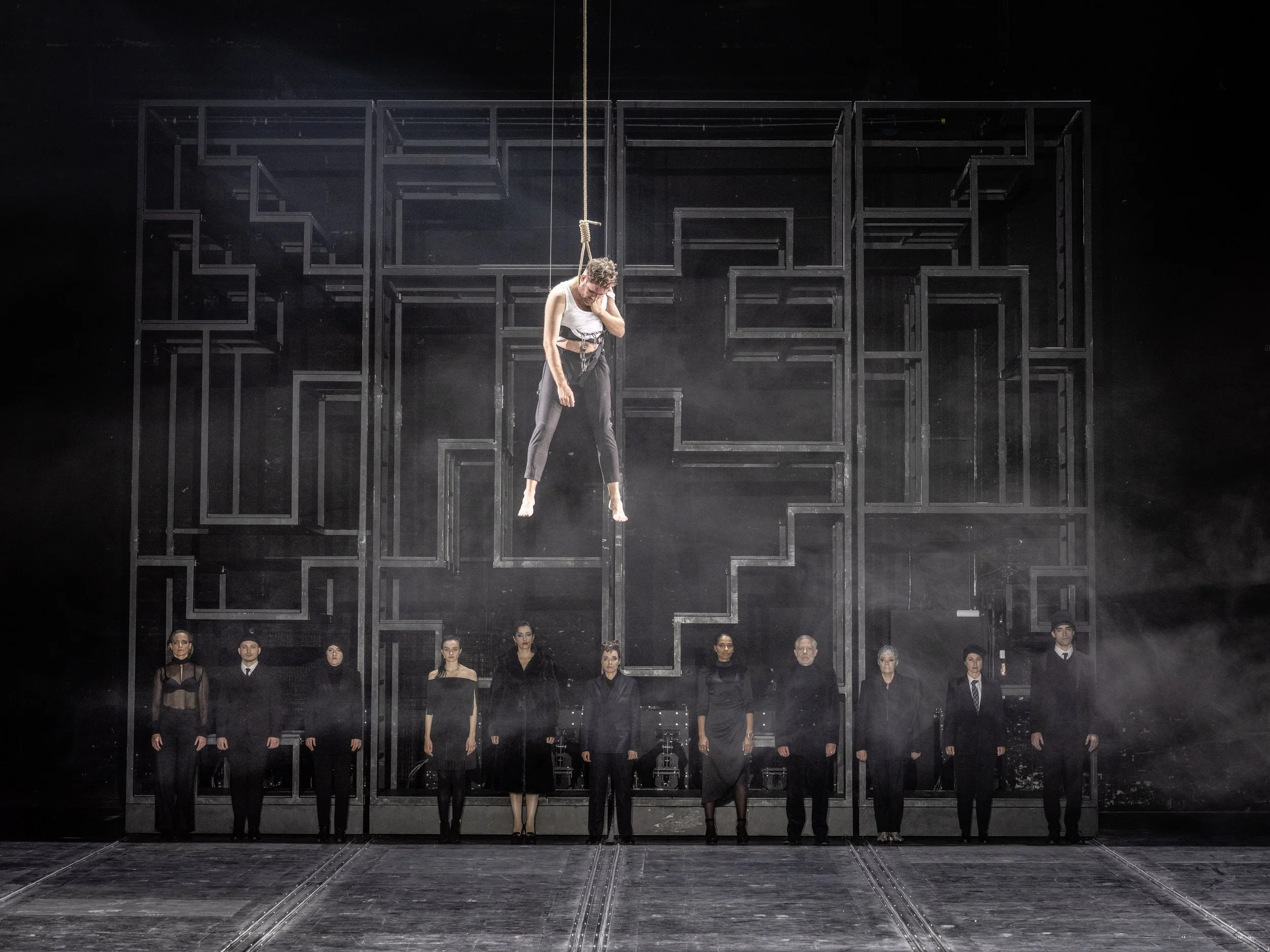 Bühnenperformance mit einer Frau, die an einem Seil hängt, vor einer schwarzen und grauen abstrakten Kulisse, mit einem linearen Muster und mehreren Menschen in schwarzen Kleidern, die im Hintergrund stehen.