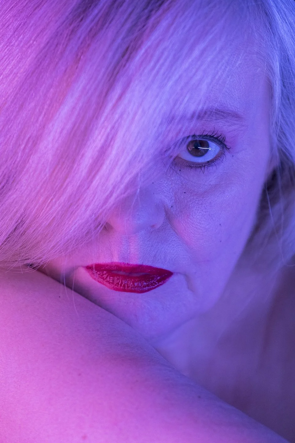 Nahaufnahme eines Gesichts mit roten Lippen, blauer Iris, Blonde Haare, teilweise verdeckt durch das Haar, unter Pink- und Blaulichtlicht.