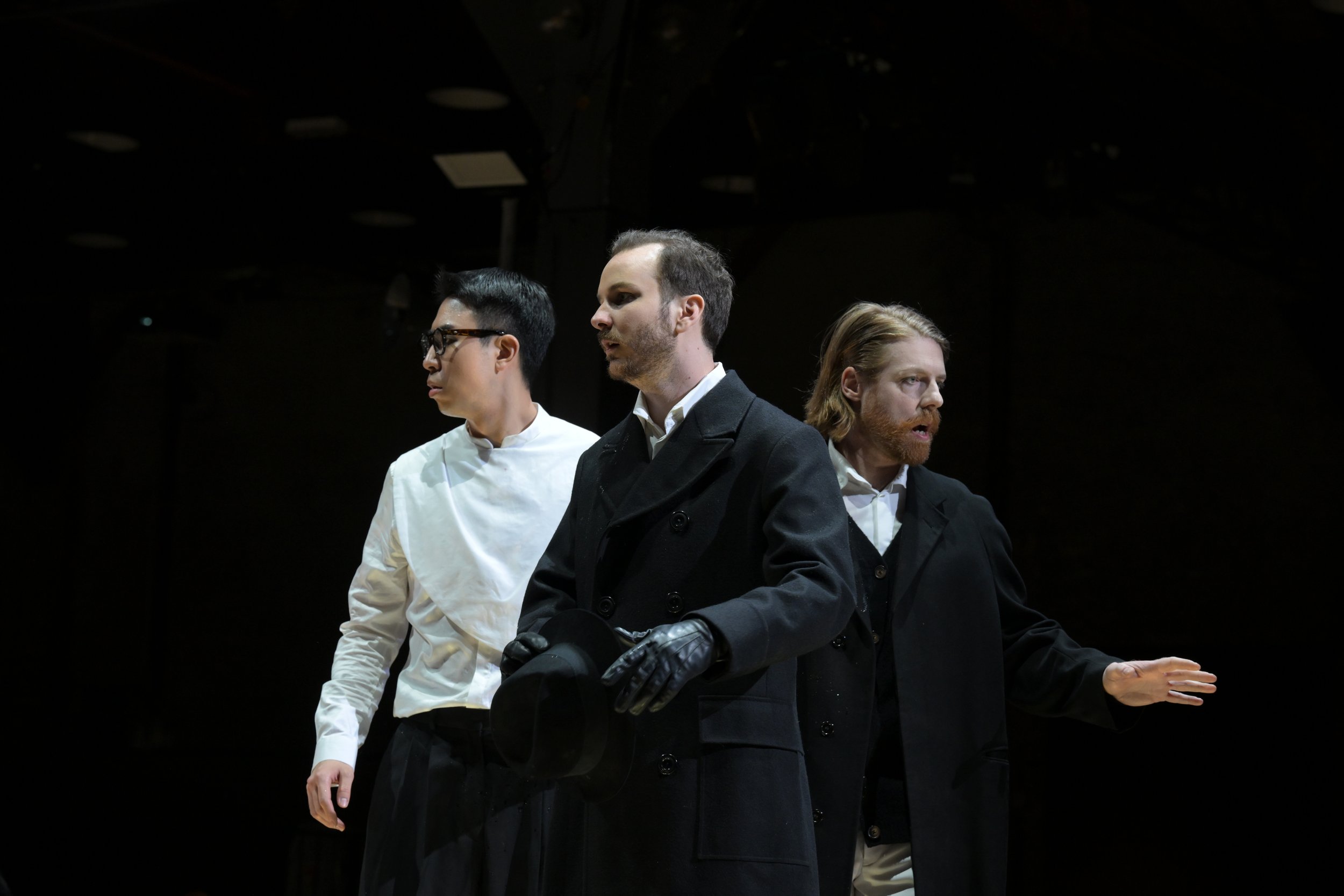 Drei Schauspieler auf einer Bühne, einer in einem weißen Hemd und Brille, der zweite in einer schwarzen Jacke und Handschuhen, der dritte in einem schwarzen Mantel, stehen in einer dramatischen Szene.