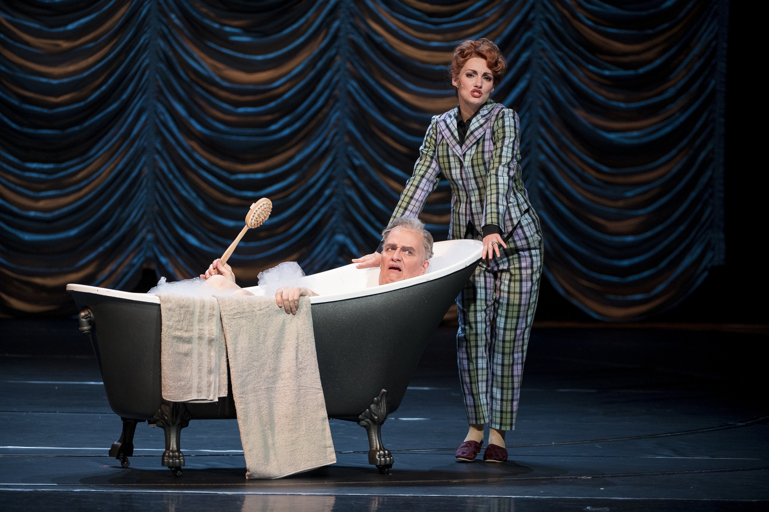 Theaterdarbietung mit einer Frau in einem Anzug neben einem Mann in einer Badewanne voller Seifenblasen, während sie auf einer Bühne vor einem dunklen, gewellten Vorhang steht.