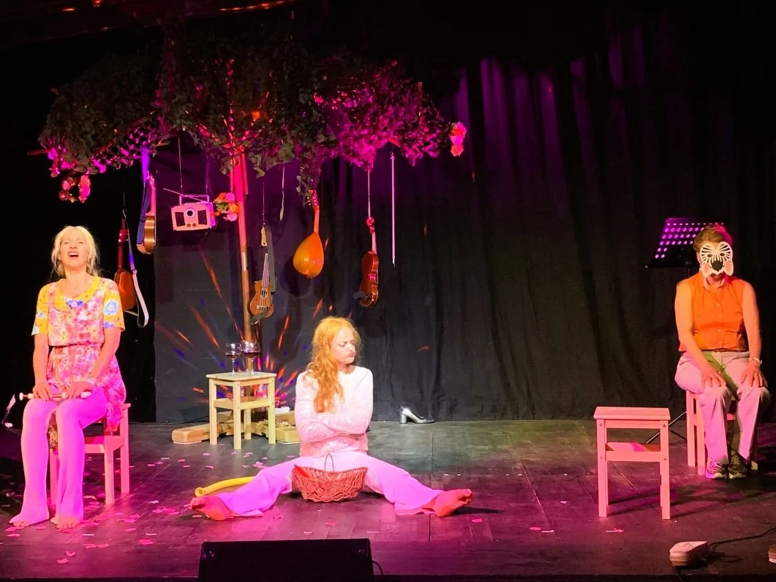 Drei Frauen auf einer Bühne, eine sitzt lachend, eine sitzt nachdenklich in der Mitte, und eine trägt eine Maske. Die Bühne ist dekoriert mit einem Baum, Musikinstrumenten und einer kleinen Tisch mit einem Radio.