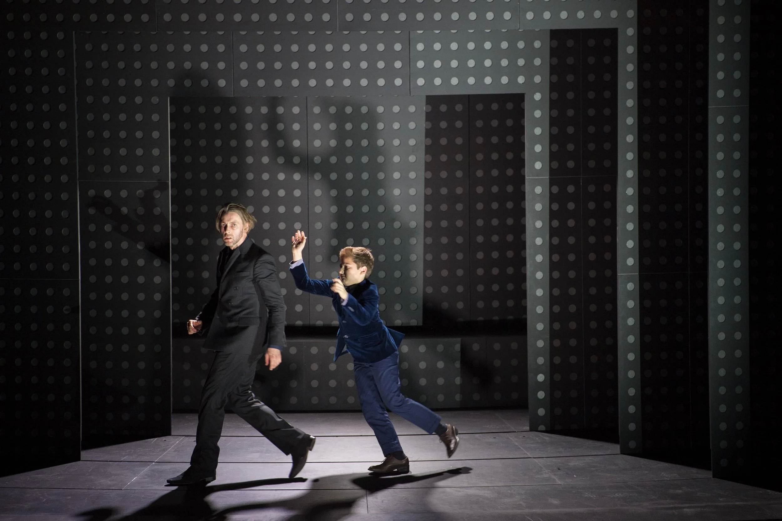 Zwei Männer in formeller Kleidung tanzen auf einer dunklen Bühne mit geometrischem, schwarz-weiß gemustertem Hintergrund.