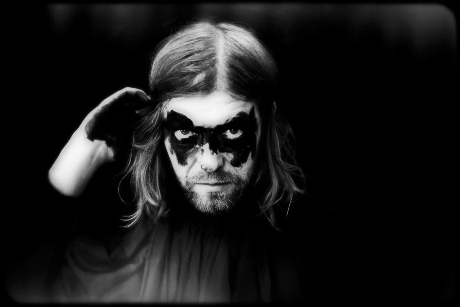 Schwarze und weiße Fotografie eines Mannes mit langem Haar und Bart, der schwarze Maske oder Make-up um die Augen trägt, blickt ernst in die Kamera, hat eine Hand an den Kopf gehoben.
