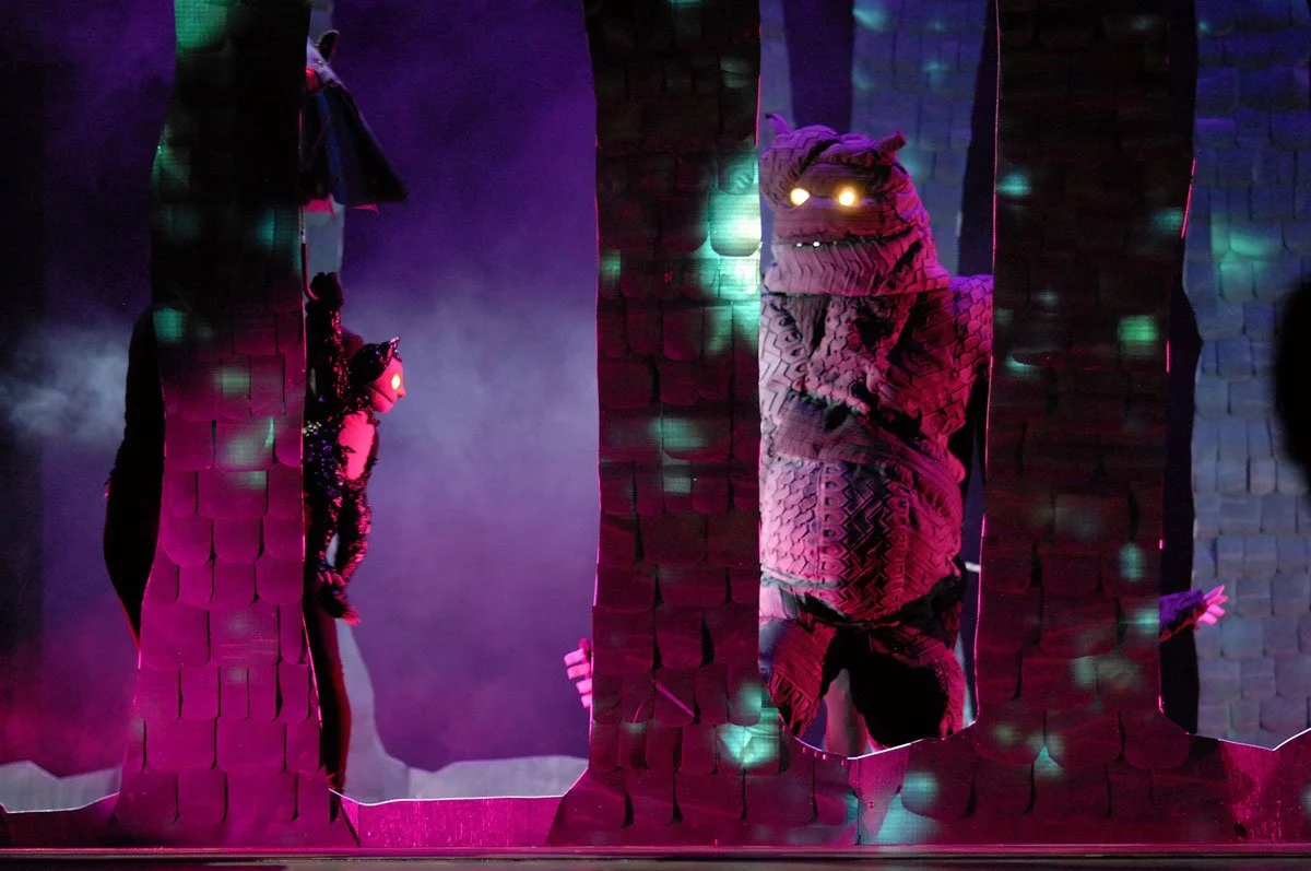 Szene mit Puppen oder Masken auf einer Theaterbühne, mit bunten Lichtern und Nebel, die einen Wald darstellen. Zwei Kreaturen mit leuchtenden Augen sind sichtbar, eine größere auf der rechten Seite und eine kleinere auf der linken Seite.