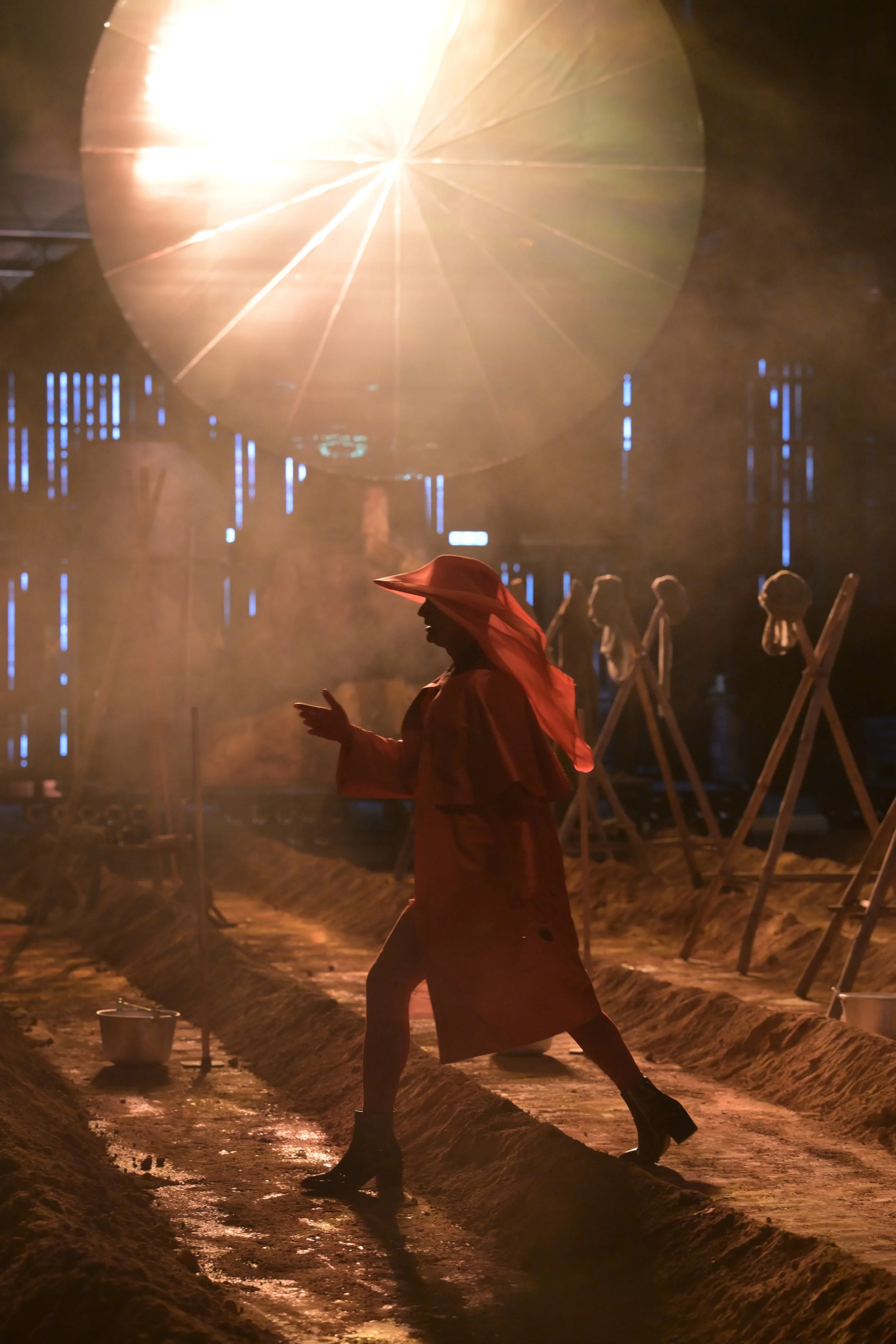 Eine Frau in rotem Kleid und Hut geht in einem dunklen, befeuerten Raum, der an ein Theaterstück oder eine Performance erinnert, mit mehreren Ständern im Hintergrund und einem großen Licht im oberen Bereich.