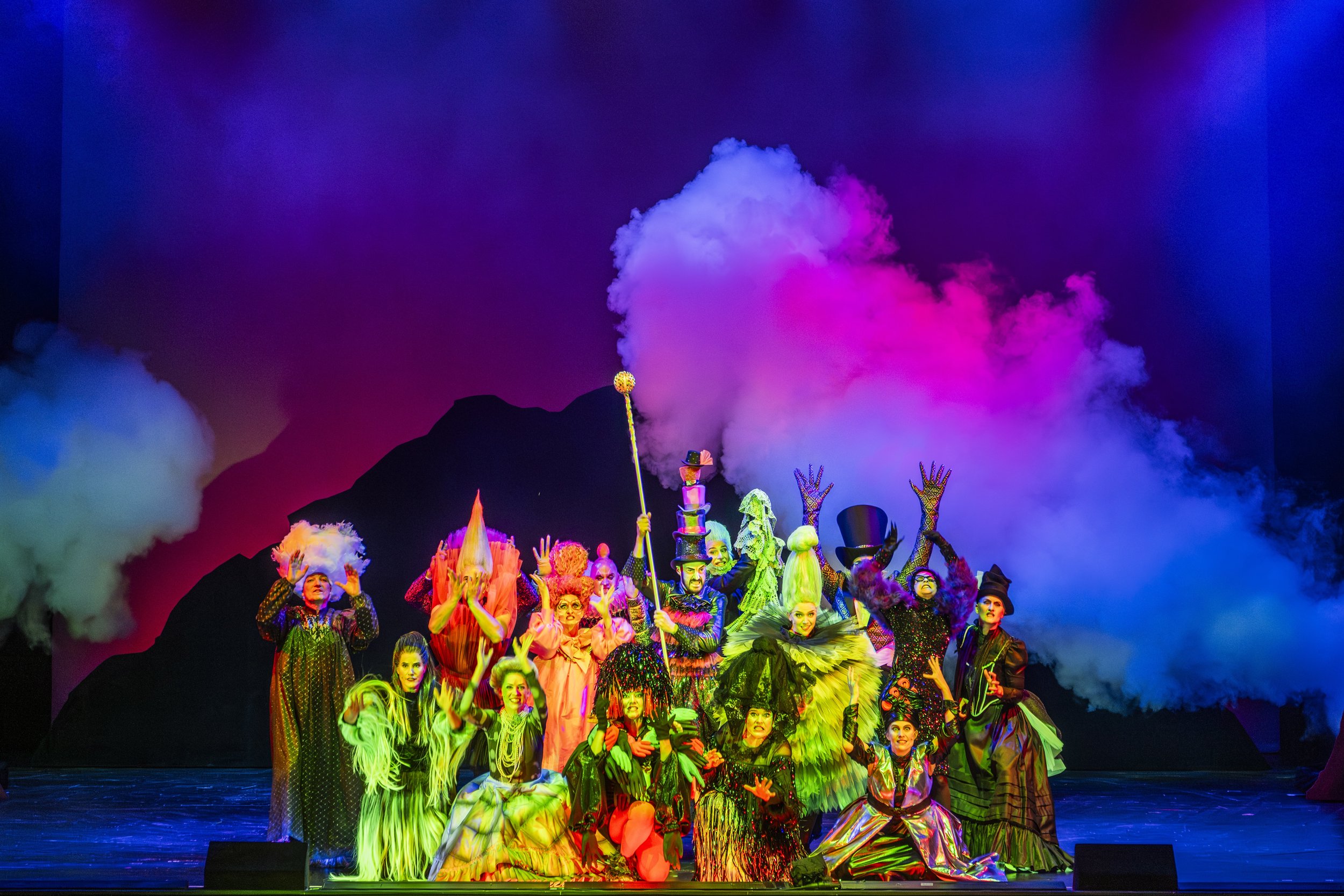 Bühnenaufnahme einer Theateraufführung mit bunten Kostümen, dramatischer Beleuchtung und Rauch dampf, die eine Fantasiewelt darstellen.