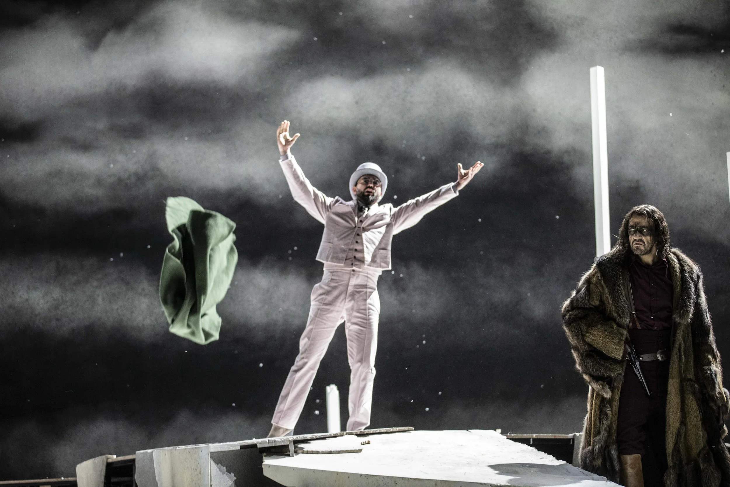 Schauspieler in Kostümen auf einer Bühne vor einem dunklen Himmel mit Wolken, einer trägt einen hellen Anzug und Hut, hebt die Arme, der andere trägt einen Pelzmantel und hat lange Haare, im Hintergrund Wolken und Nebel.