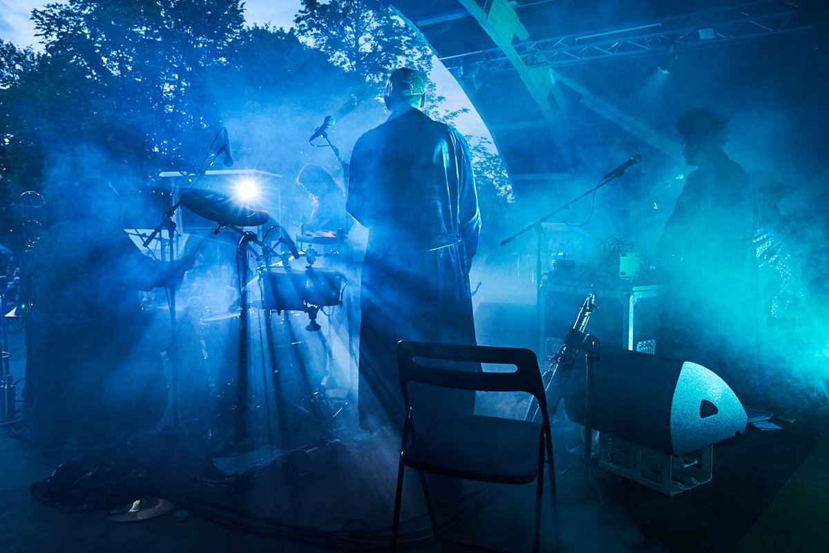 Musiker auf einer Bühne im Dämmerlicht, umgeben von blauer Nebel, mit Musikern und Instrumenten im Hintergrund.