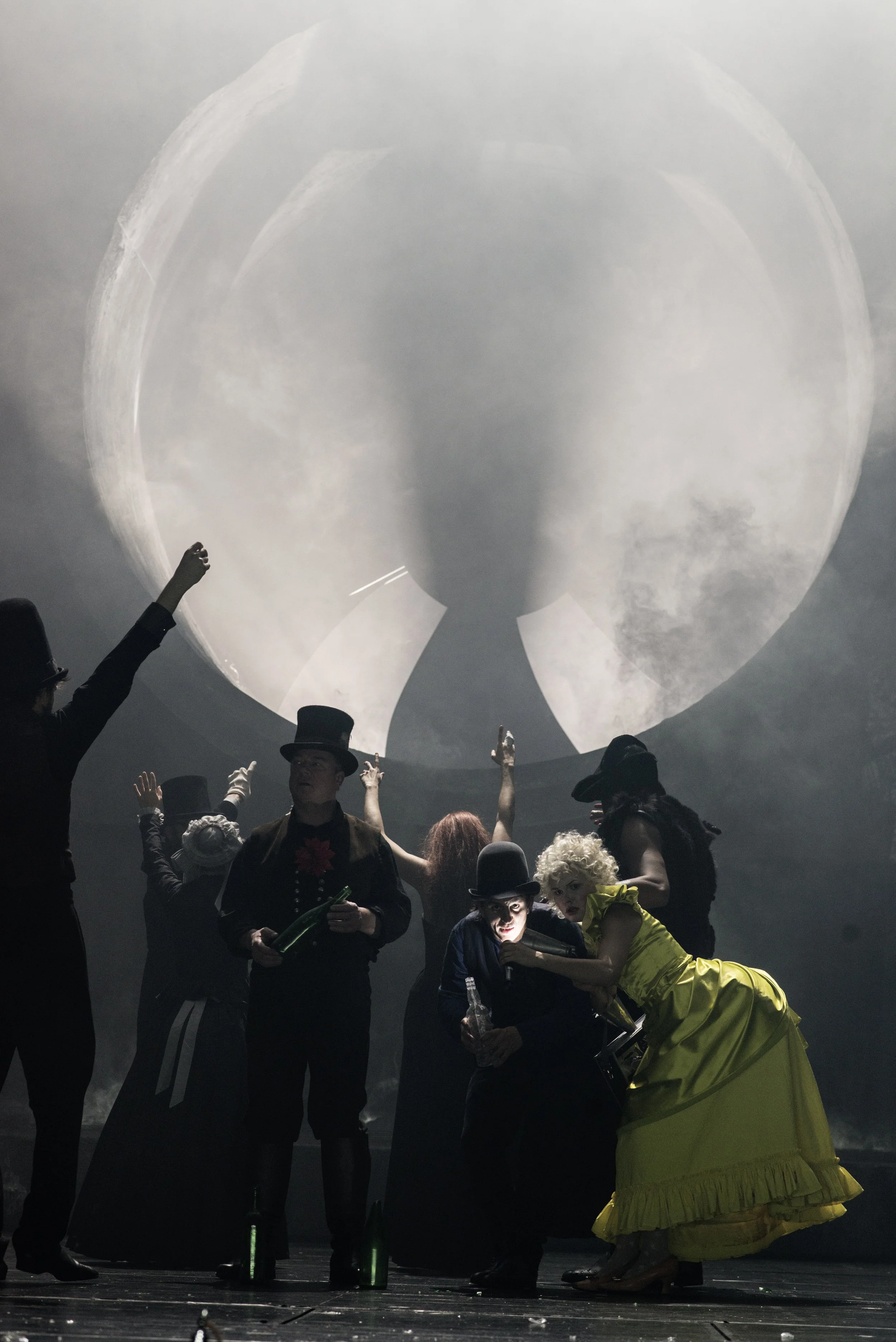 Bühnenbild mit mehreren Menschen in Kostümen, einer großen Mondprojektion im Hintergrund.
