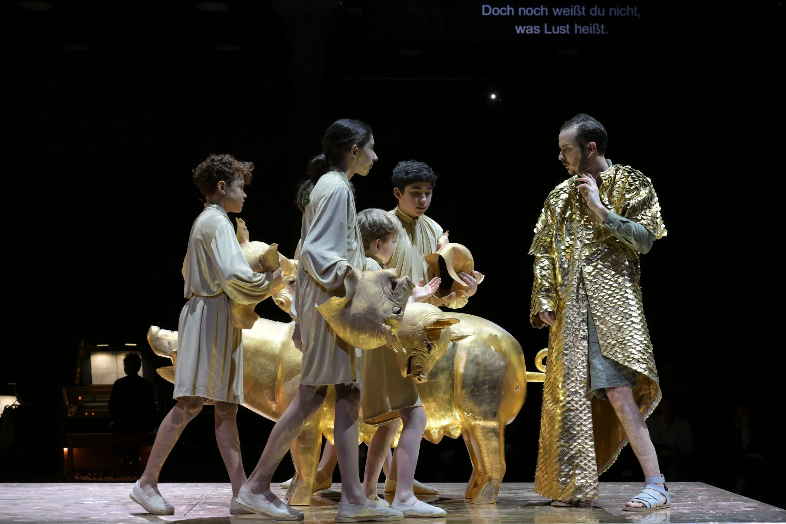 Szenenfoto aus einer Theaterproduktion mit fünf Kindern in weißen Kostümen um einen goldfarbenen Elefanten aus Metall, ein Erwachsener in einer goldfarbenen Schuppenjacke steht vor ihnen. Im Hintergrund ist eine Projektion mit deutschem Text zu sehen