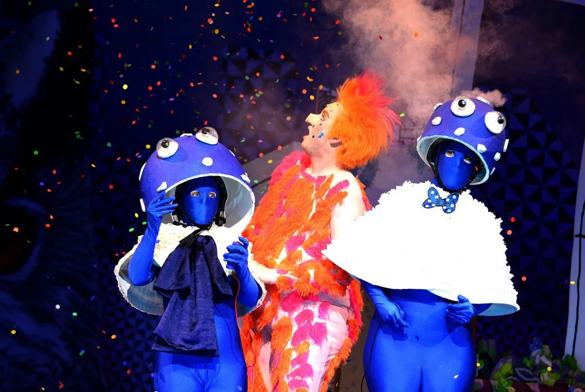 Drei Personen in bunten Kostümen, zwei in blauen Anzügen mit Pilzkoepfen, und eine in einem pink-orange Fellkostüm, bei einer Aufführung mit Konfetti im Hintergrund.
