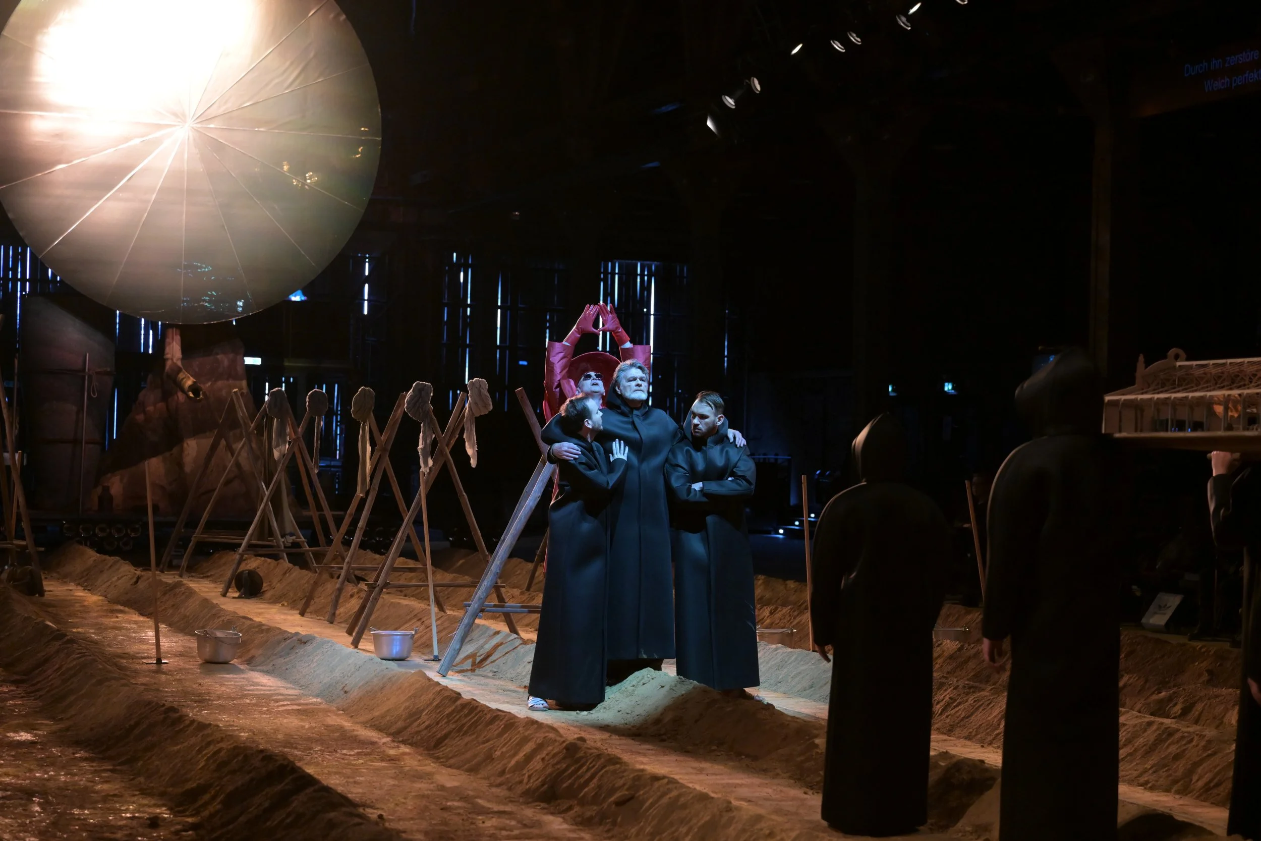 Ein Bühnenbild mit drei Menschen in schwarzen Roben, die in einer Szene aus einem Theaterstück oder einer Oper stehen. Im Hintergrund sind Requisiten wie Schaufeln und Eimer, die eine Baustellenszene darstellen, sowie eine große Beleuchtung. Die Stim