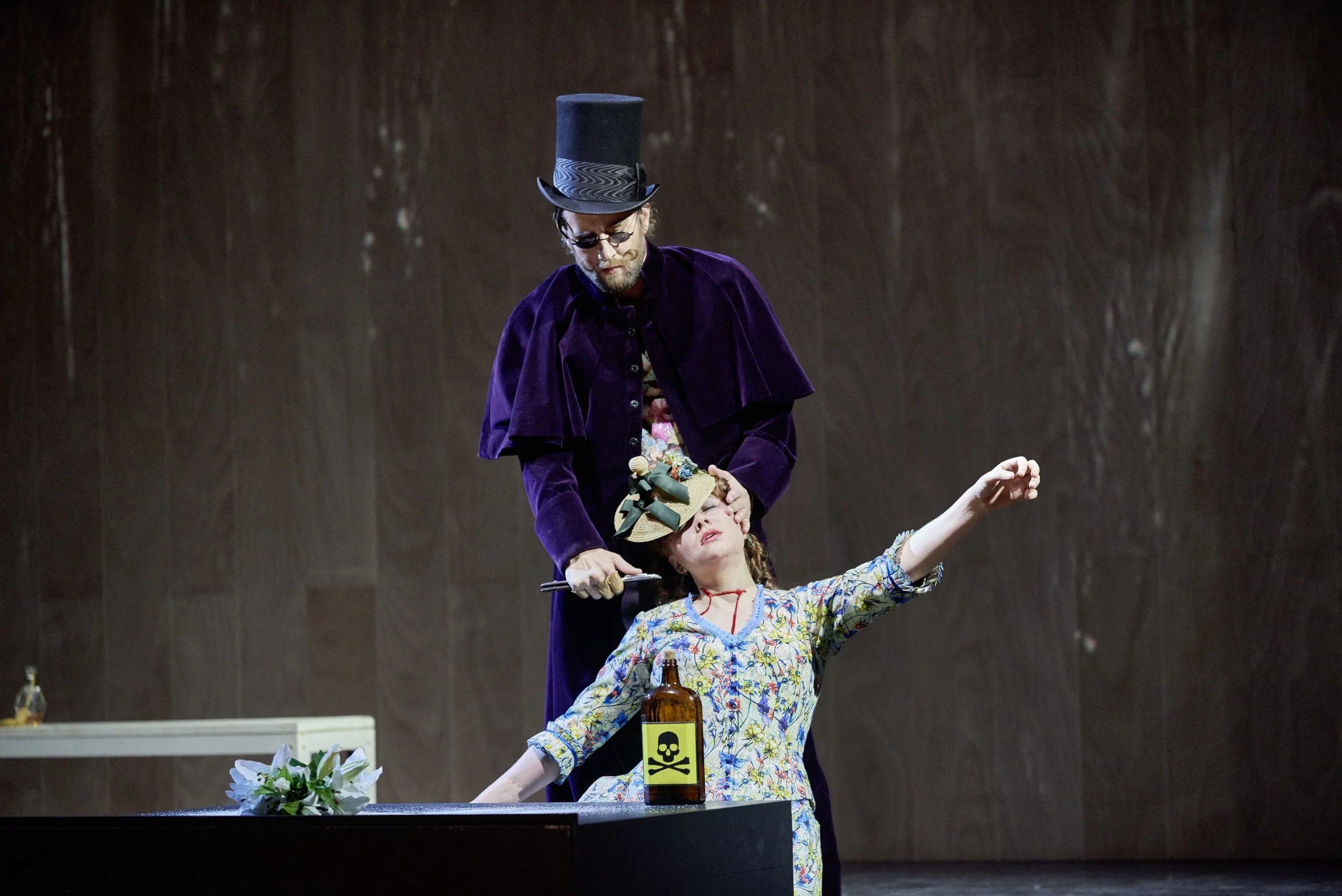 Theaterstück mit zwei Schauspielern, einer männlich im lila Mantel und Zylinder, der andere weiblich im bunten Kleid mit Hut, Szene am Tisch mit verseuchter Flasche und Blumen.