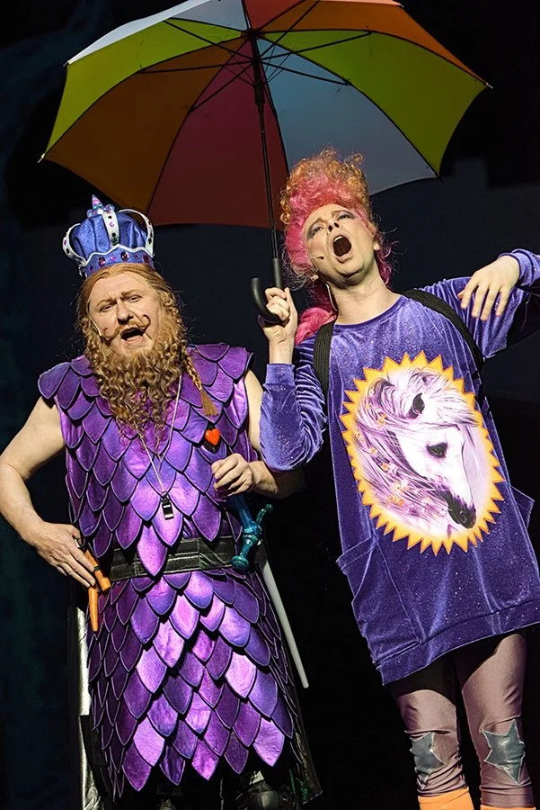 Zwei Schauspieler in fantasievollen Kostümen, einer trägt eine Krone und lila Schuppen, der andere hat pink gefärbtes Haar, trägt ein T-Shirt mit einem Einhorn-Design, hält einen bunten Regenschirm und singt auf einer Bühne.