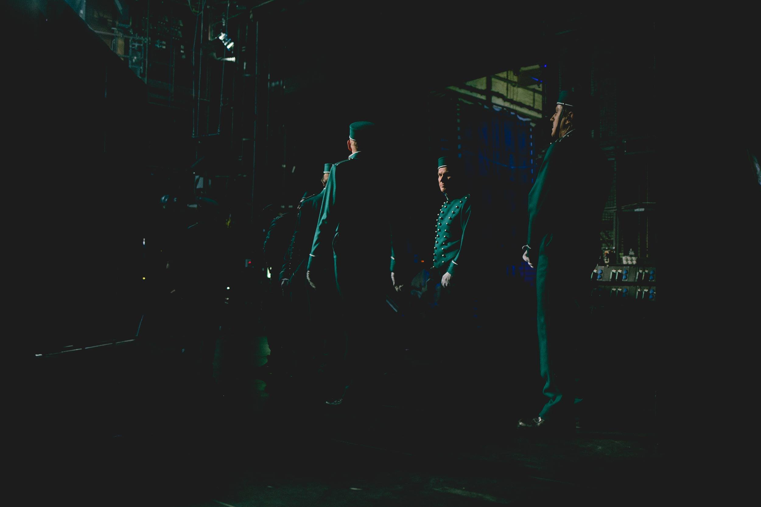 Mehrere Männer in formeller Kleidung stehen in einer dunklen Umgebung, was auf eine Szene im Theater oder eine Zeremonie hindeuten könnte.