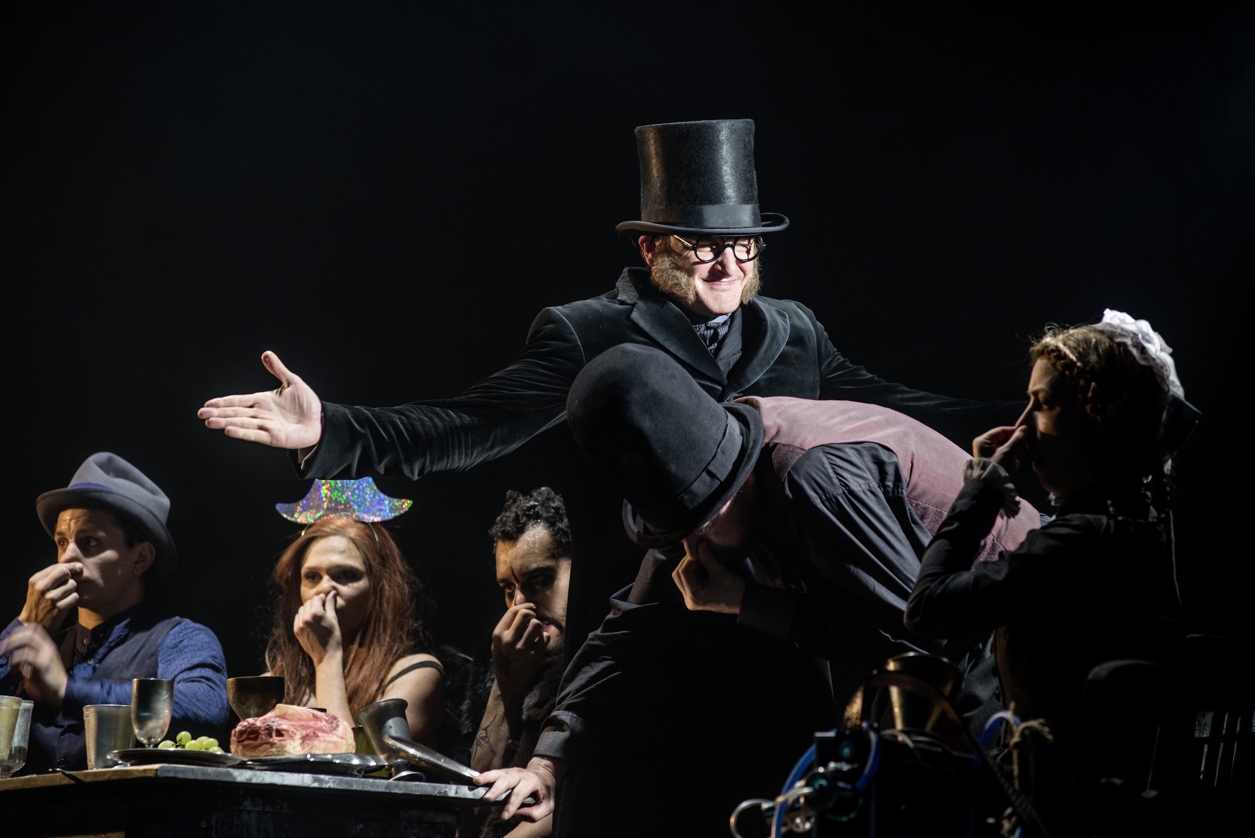 Szenische Darstellung einer Theatergruppe mit Menschen in Kostümen an einem Tisch, einer trägt einen Zylinderhut, andere tragen Hüte und haben verschiedene Posen. Die Szene ist dunkel gehalten, mit Fokus auf die Akteure.