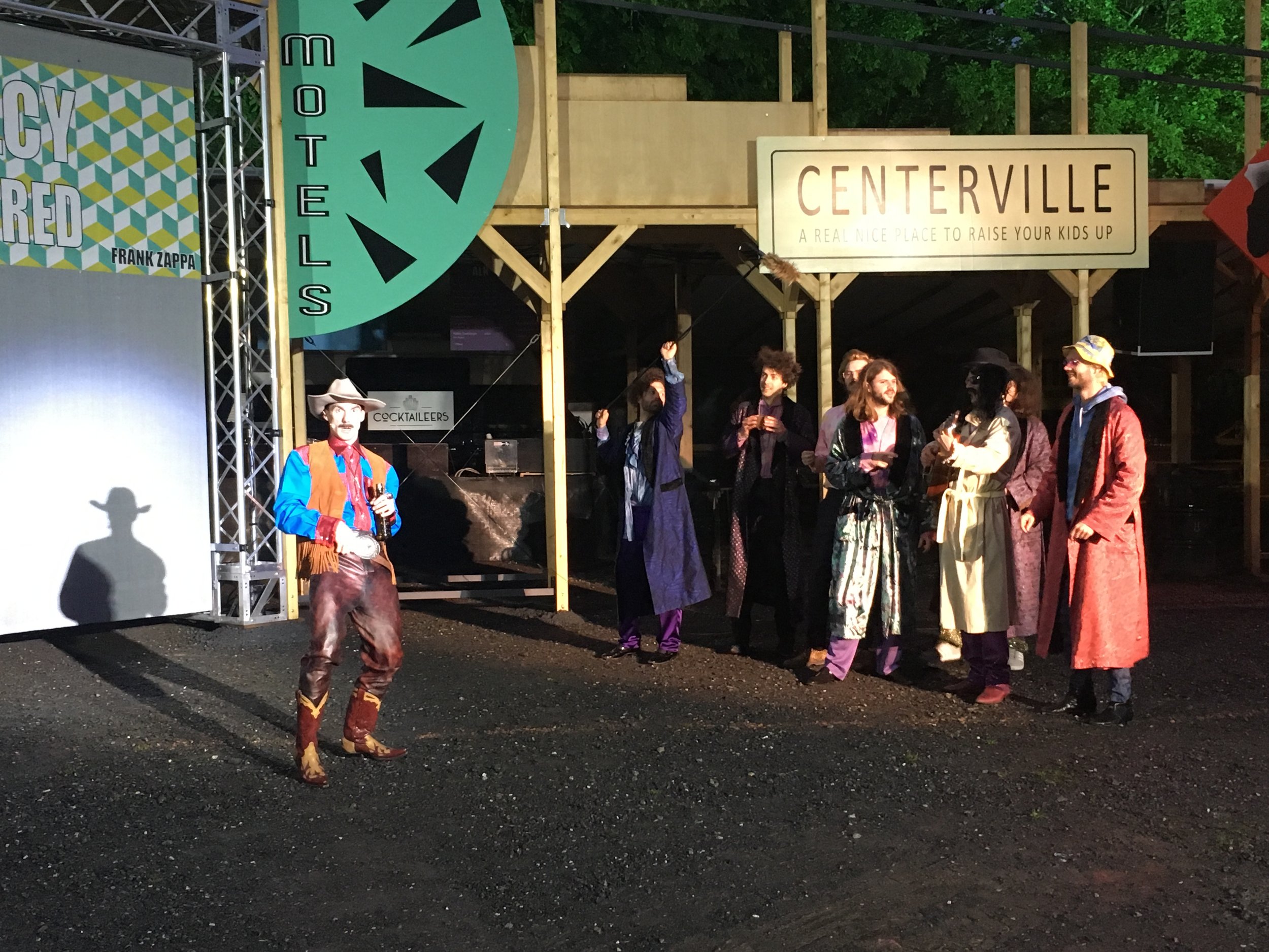 Eine Gruppe von Menschen in bunten, historischen Kostümen steht vor einem Holzständer bei einem Event. Es gibt ein Schild mit der Aufschrift 'CENTERVILLE' und einen weiteren mit 'HOTEL'. Einer der Männer trägt einen Cowboy-Hut und ein rotes Weste, wä