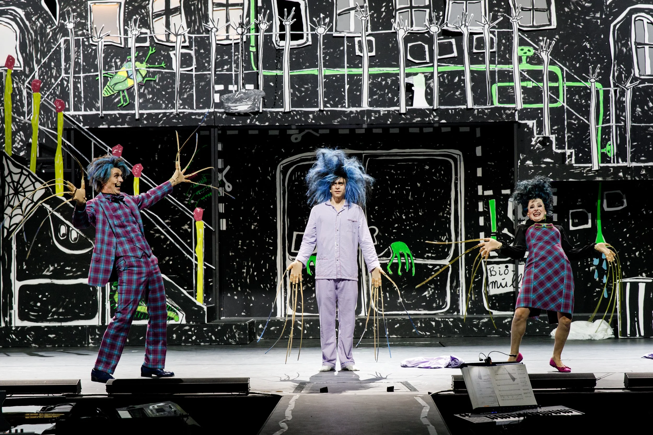 Drei Darsteller in bunter Kleidung und surrealen Frisuren auf einer Theaterbühne mit schwarzem und weißen Hintergrund bemalt, was eine Hausfassade darstellt. Die Darsteller wirken in einer aktiven, clownartigen Performance.