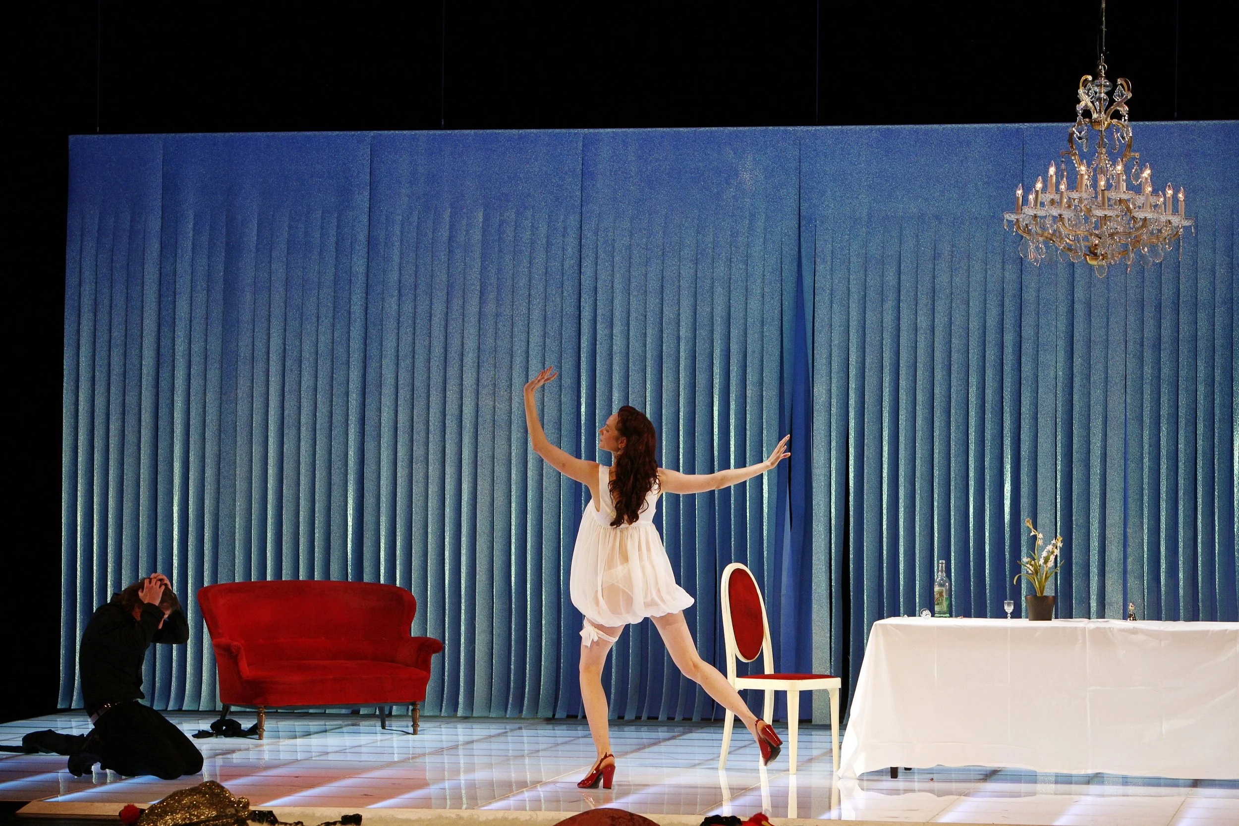 Eine Bühne mit blauer Vorhang, eine Tänzerin in weißem Kleid und roten Schuhen tanzt, während ein Fotograf sie fotografiert. Es gibt einen roten Sessel, einen weißen Tisch mit einer Vase und einer Flasche, und einen eleganten Kronleuchter.