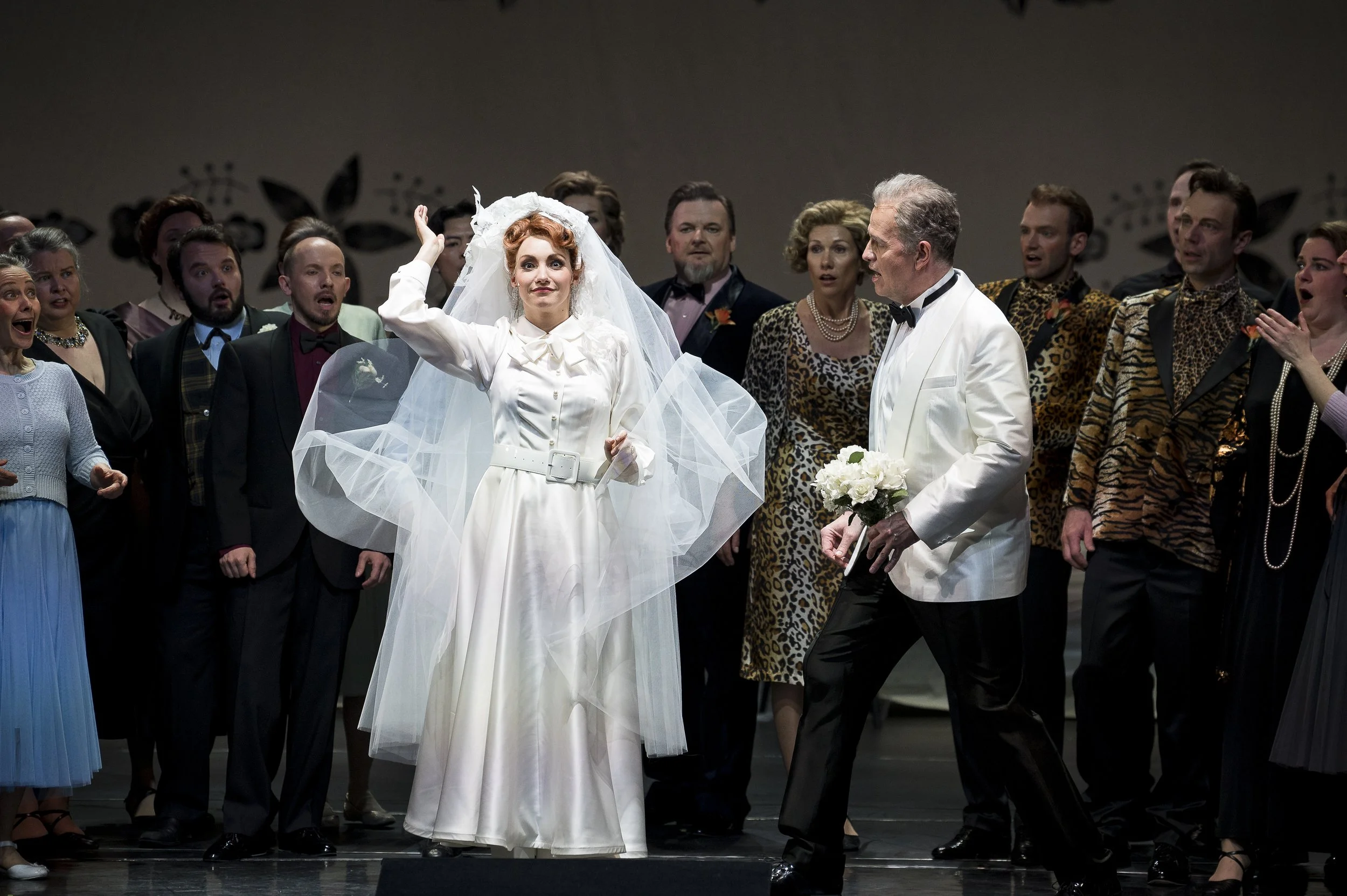 Bühnenfoto einer Theaterproduktion mit mehreren Darstellern, darunter eine Frau in einem weißen Brautkleid mit Schleier und ein Mann in weißem Anzug mit Blumen, umgeben von anderen Schauspielern in bunten Kostümen.